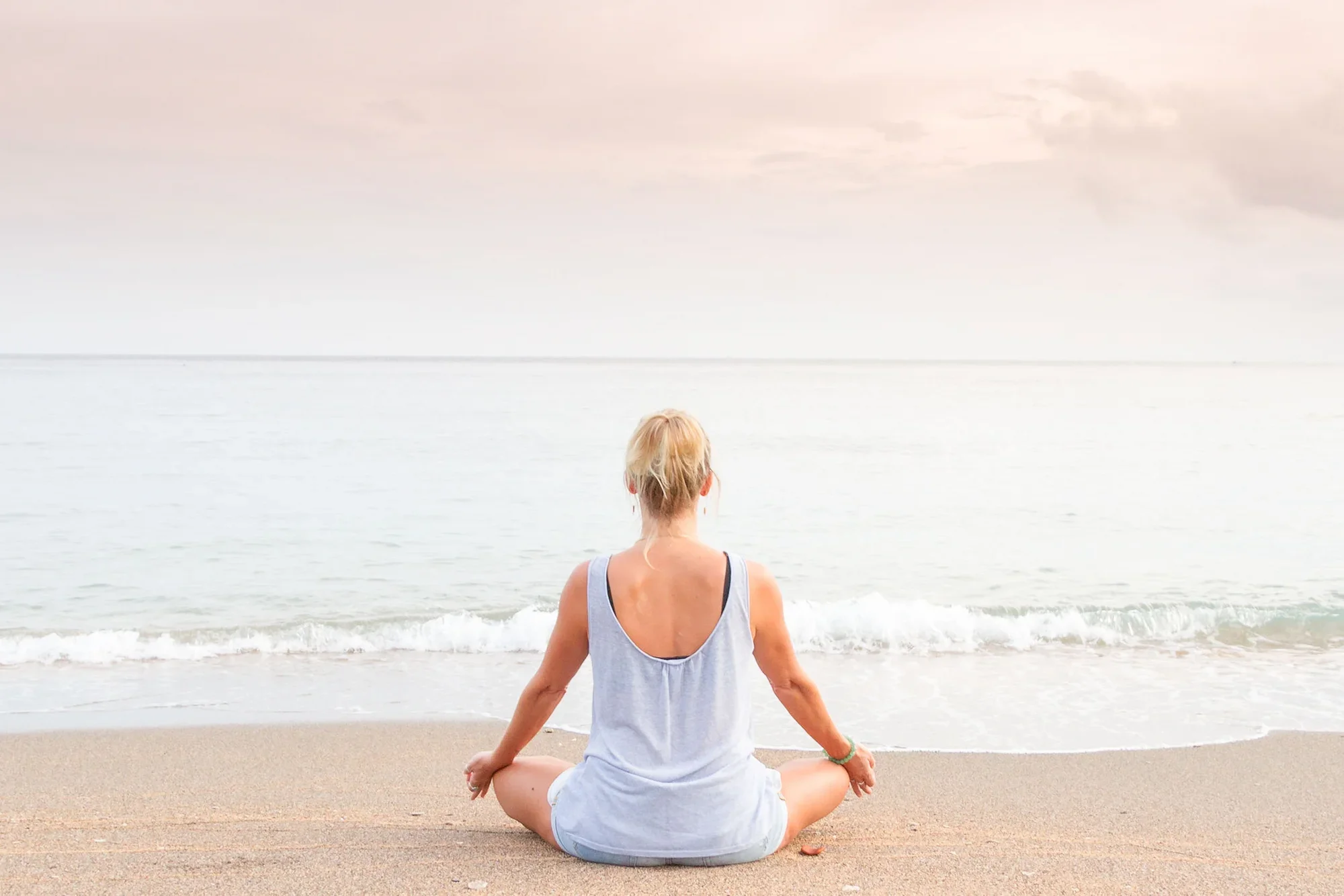 Back view of a blonde woman sitting cross-legged in a peaceful lotus meditation pose on a sandy beach shore, looking out towards gentle ocean waves under a soft pastel sunset sky.