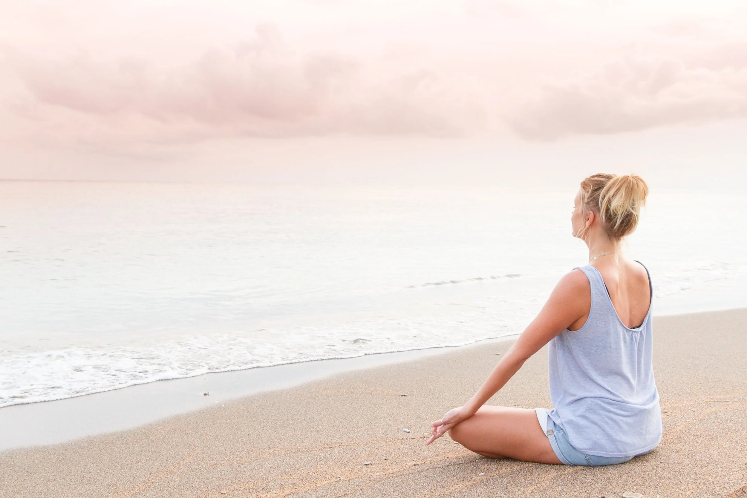 Profile view of a blonde woman sitting cross-legged in meditation on a sandy shore facing gentle ocean waves under a soft pastel pink sky, photographed by Seña Studio.