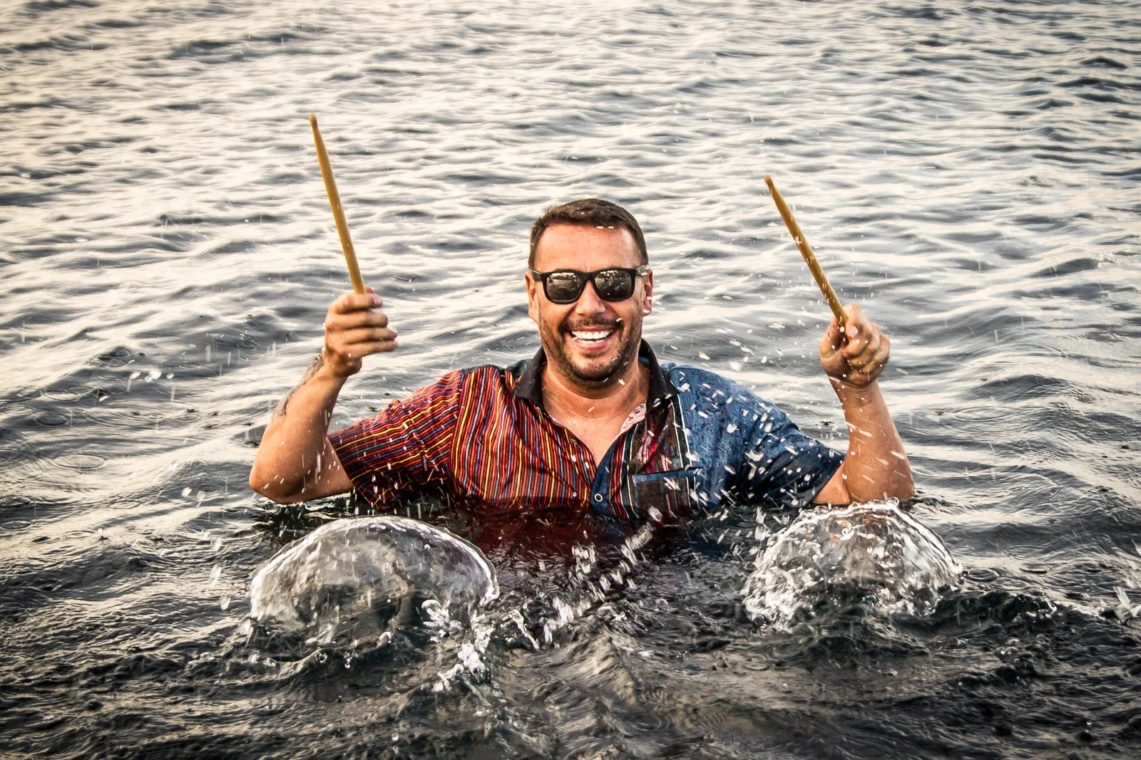 Action portrait of a smiling male drummer wearing sunglasses wading chest-deep in ocean water, raising his drumsticks as water splashes around him, photographed by Seña Studio.