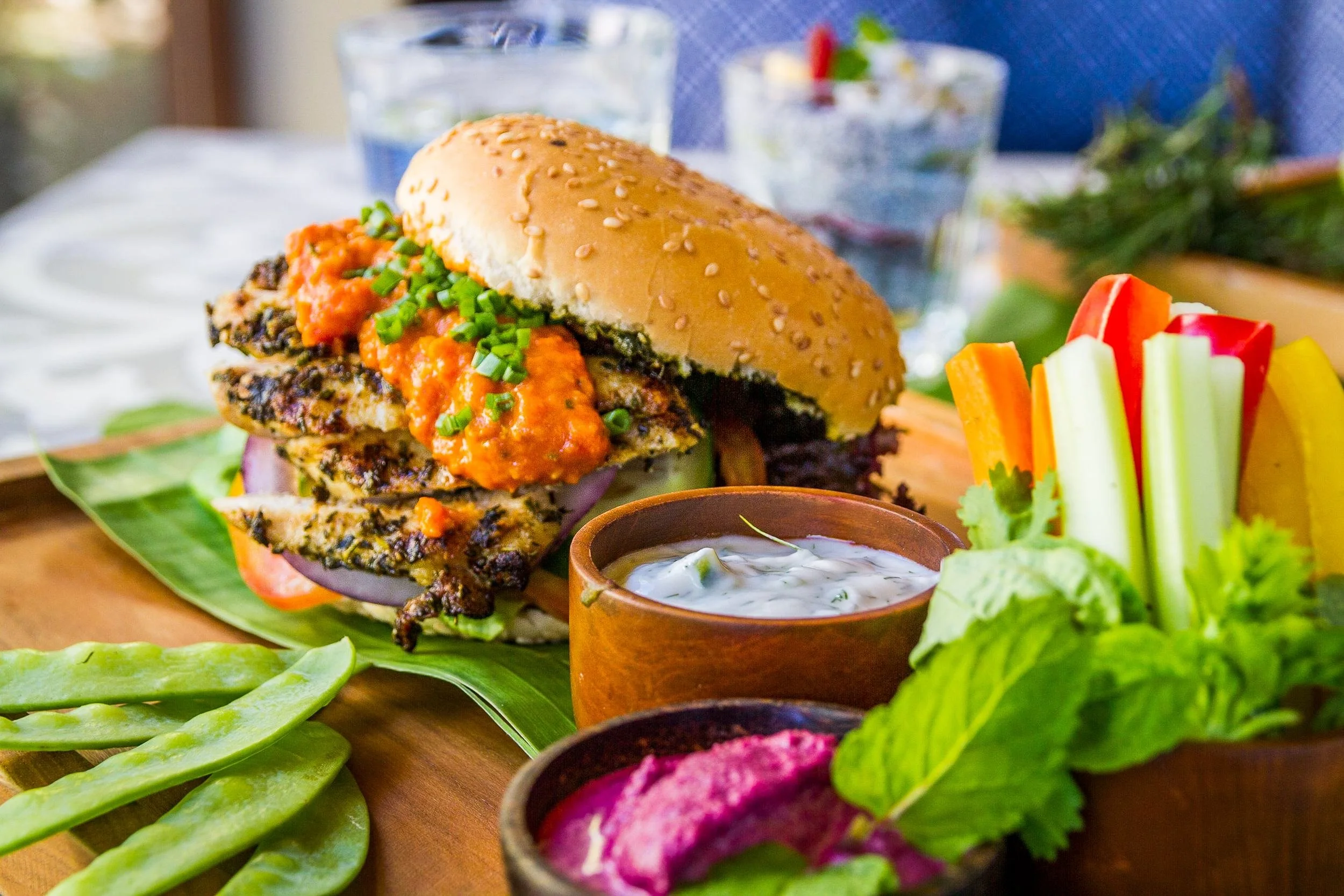 Close-up food photography of a gourmet grilled vegan burger topped with savory red sauce, served on a banana leaf platter alongside fresh vegetable sticks and dipping sauces, photographed by Seña Studio.