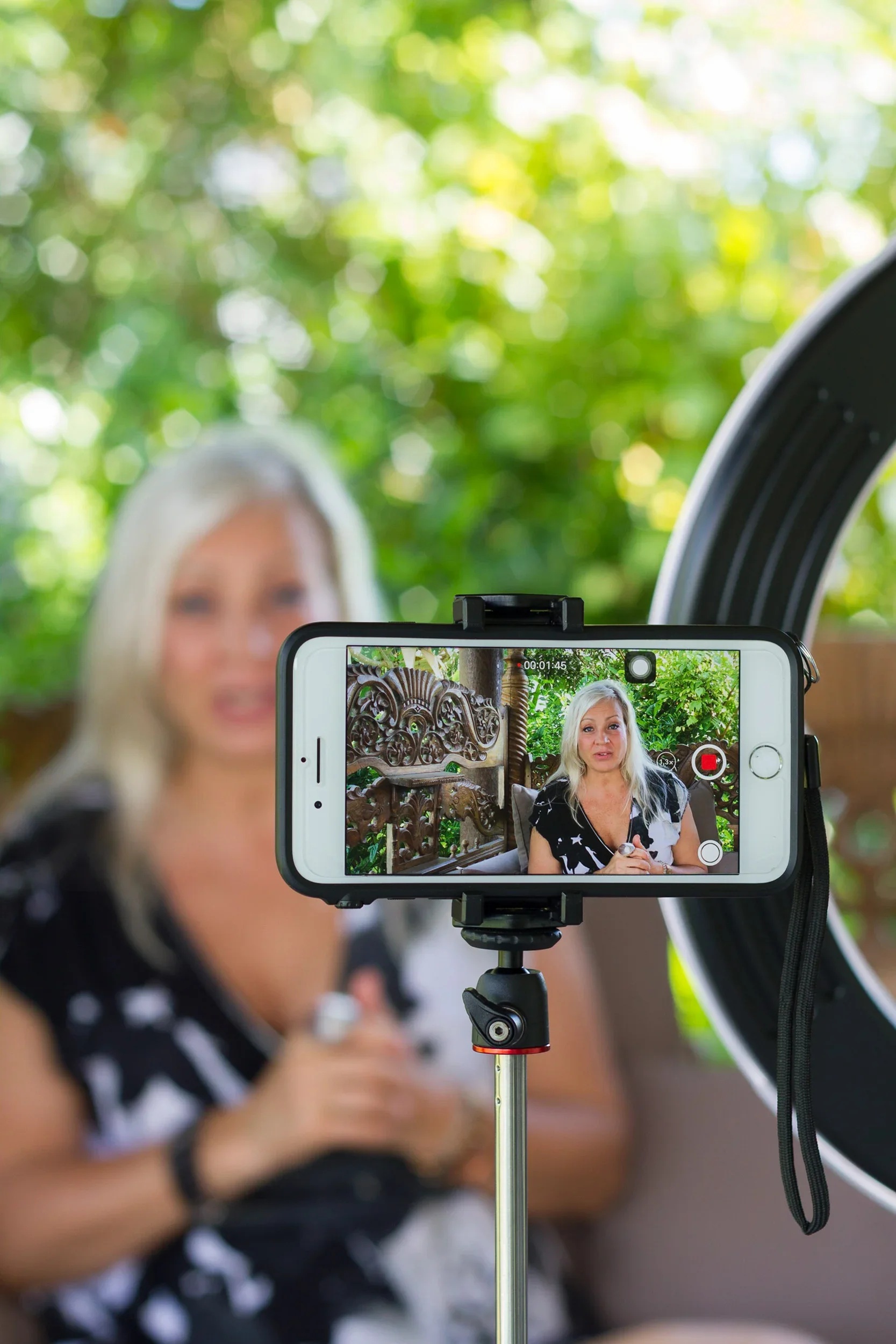 Smartphone on a tripod setup with a ring light recording a video of a blonde woman speaking.