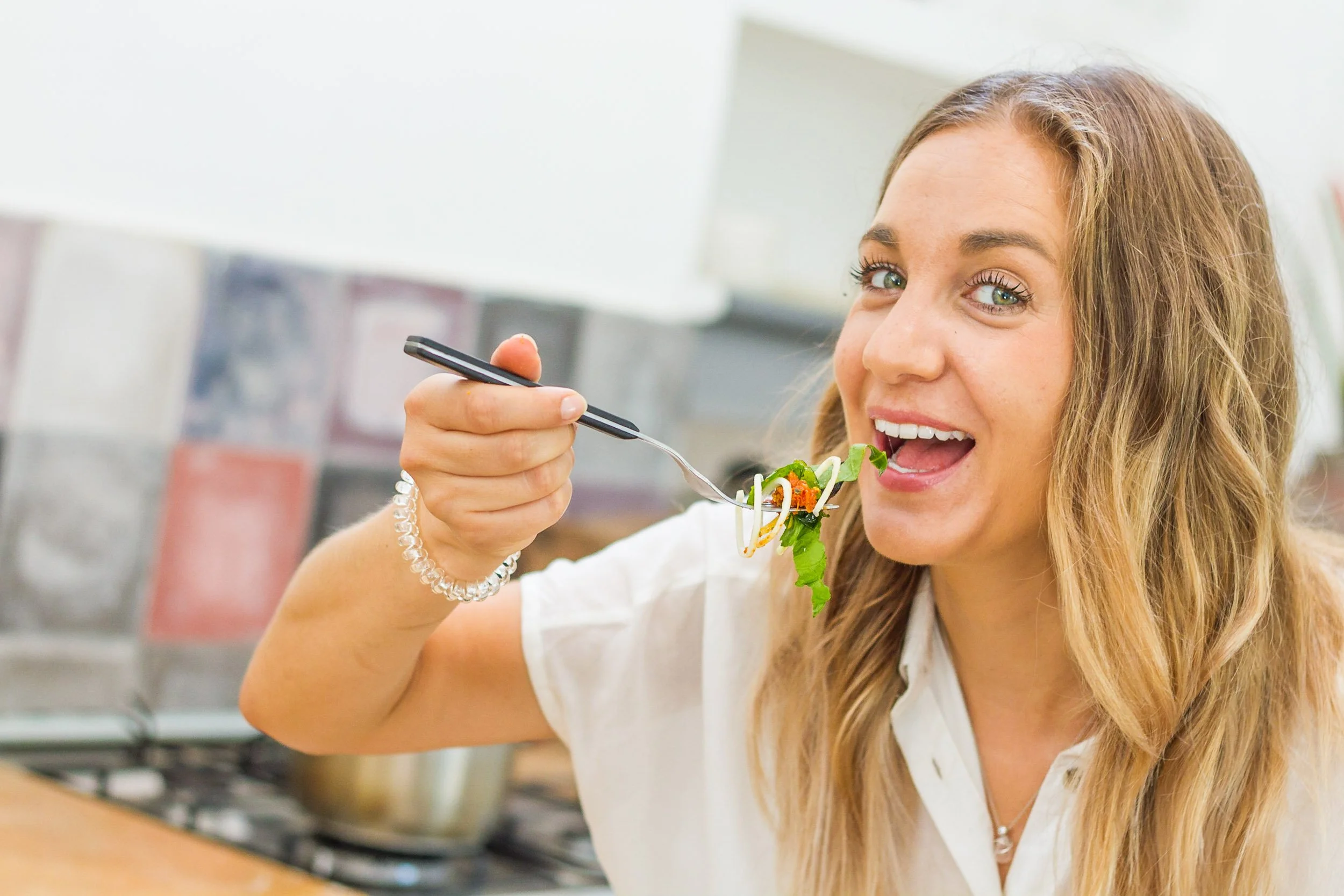 A smiling woman holding up a forkful of fresh zucchini noodles and salad close to the camera in a kitchen setting.