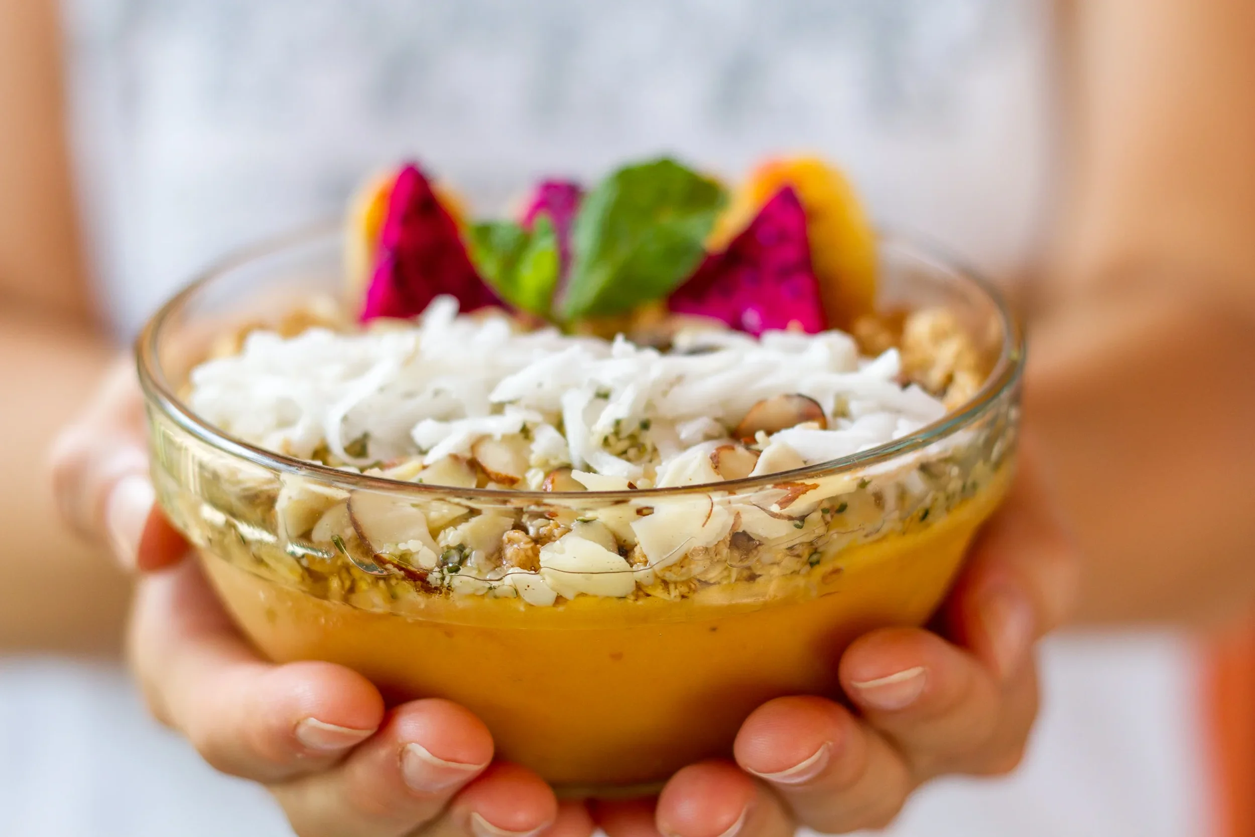 Close-up shot of a nutritious mango and peach smoothie bowl held in two hands, topped with granola, sliced dragon fruit, and shredded coconut, photographed by Seña Studio for hospitality branding.