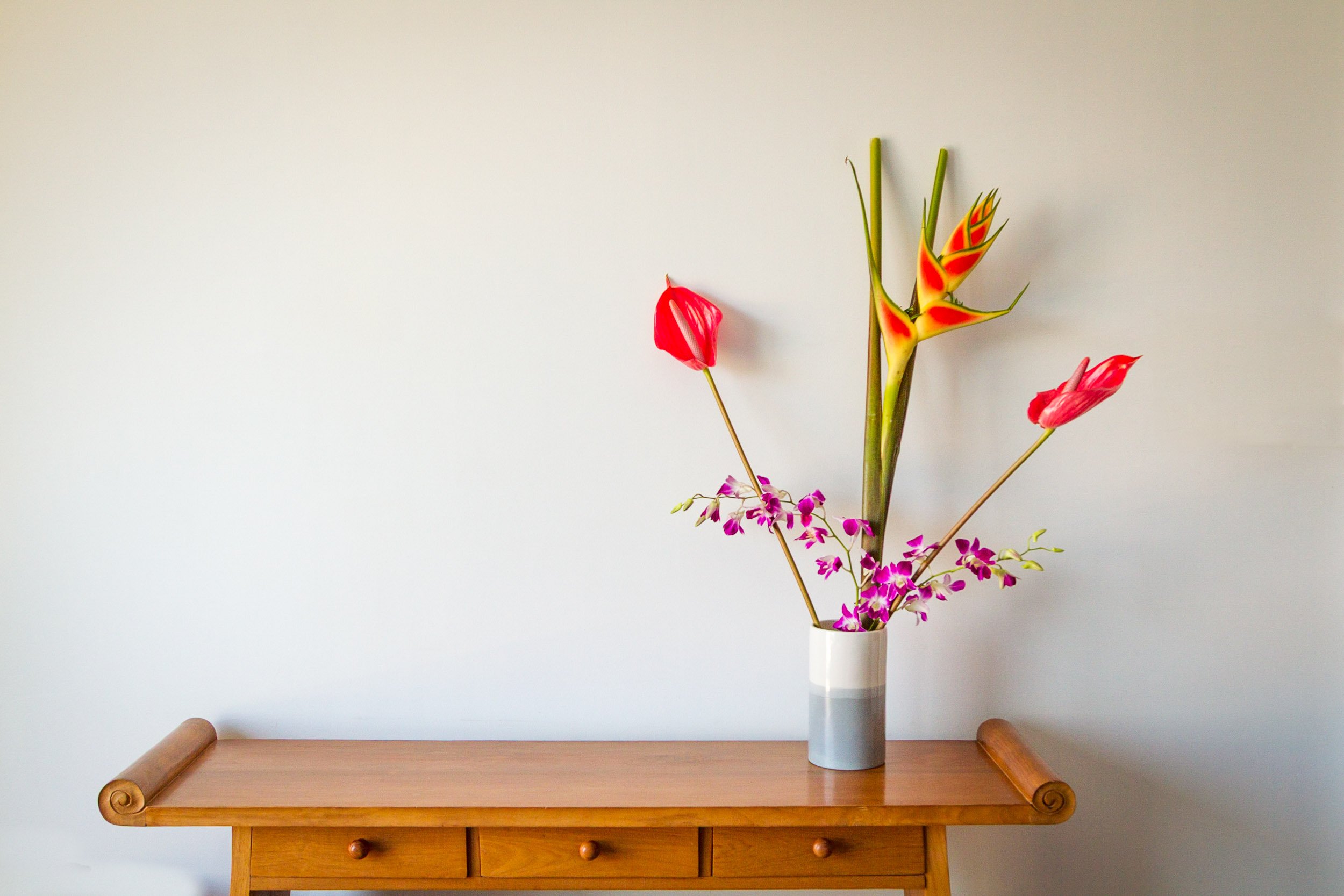 Minimalist interior decor featuring a ceramic vase with vibrant red anthuriums, heliconia, and purple orchids placed on a warm wooden console table against a neutral wall.