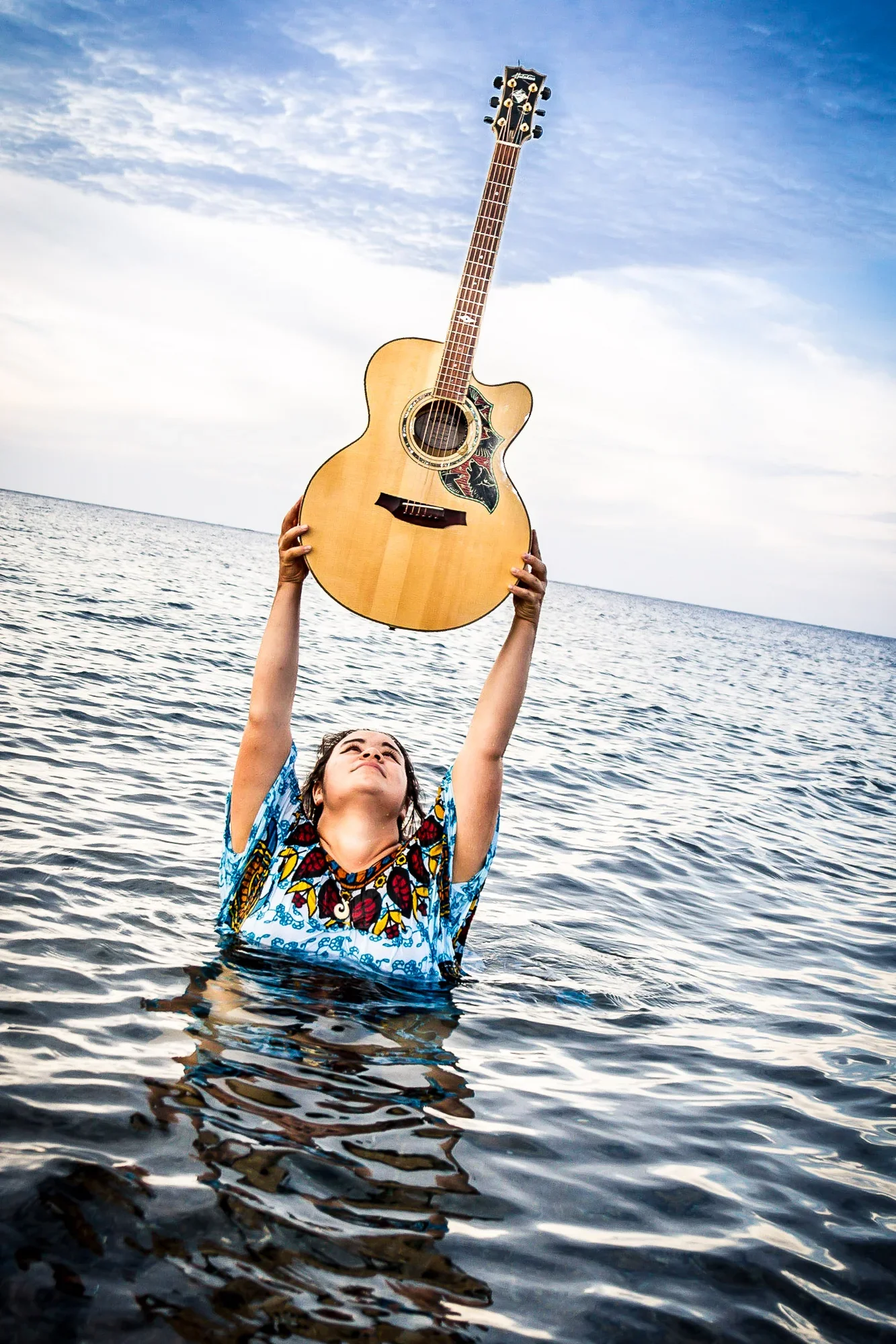Creative portrait of a female singer, Lucy Dobrovodska, wading in ocean water while lifting her acoustic guitar high above the sea surface under a bright sky, photographed by Seña Studio.