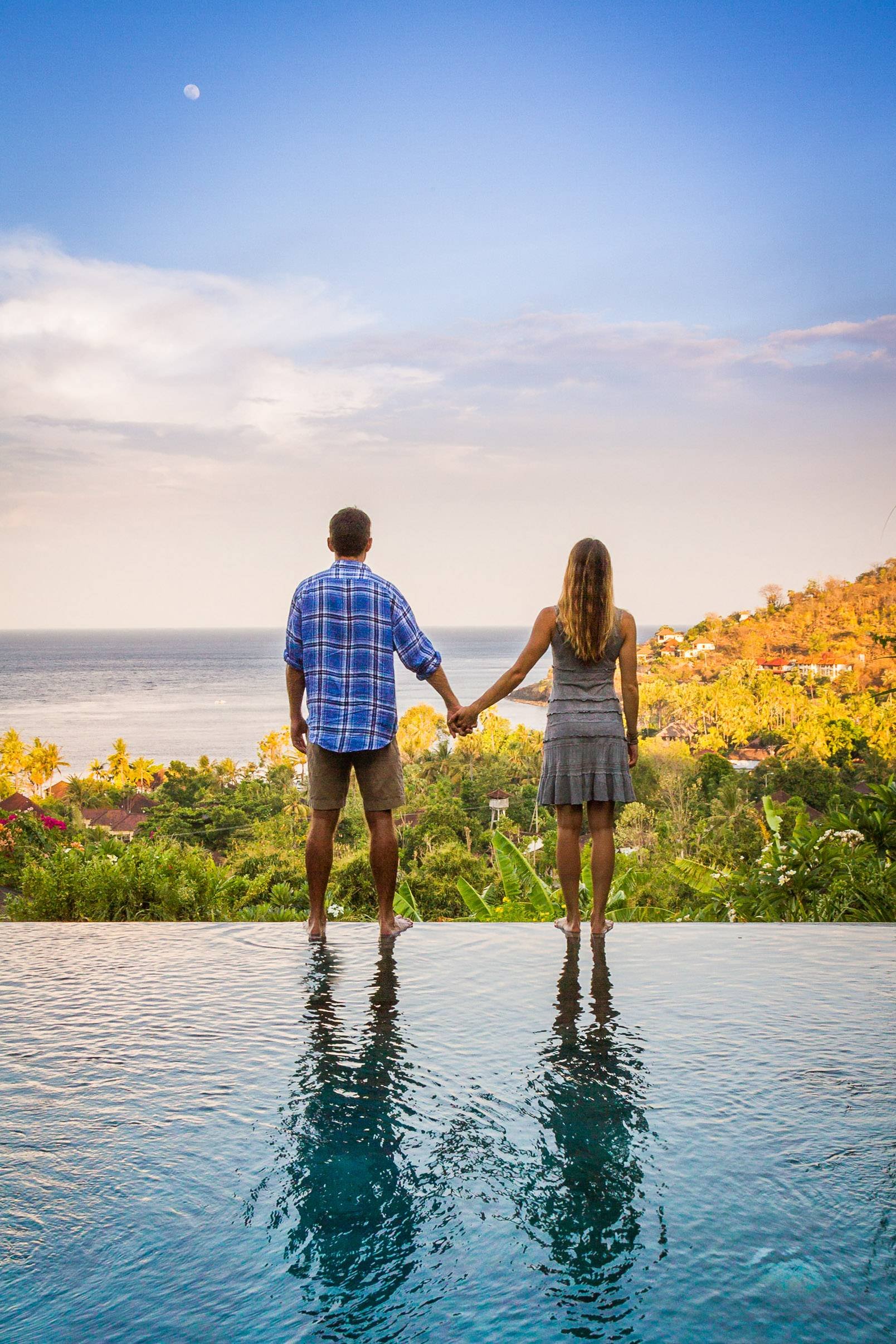 Back view of a couple holding hands while standing on the wet edge of an infinity pool with clear water reflections, looking out over a tropical ocean bay and coastal hills at dusk, photographed by Seña Studio. Photo taken at The Griya Villas & Spa, 