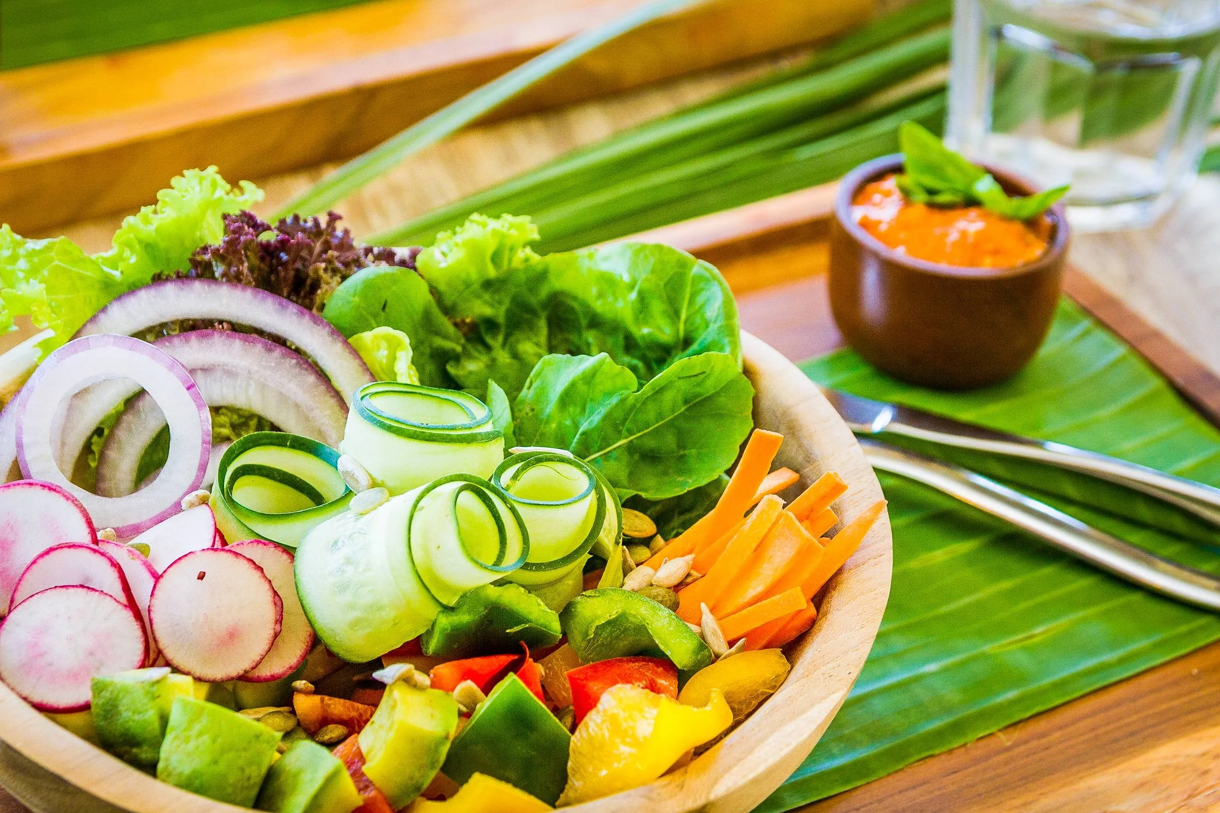 Close-up food photography of a wooden bowl filled with a colorful garden salad featuring cucumber ribbons, sliced radishes, red onion rings, diced avocado, and fresh greens with a side dipping sauce, photographed by Seña Studio.