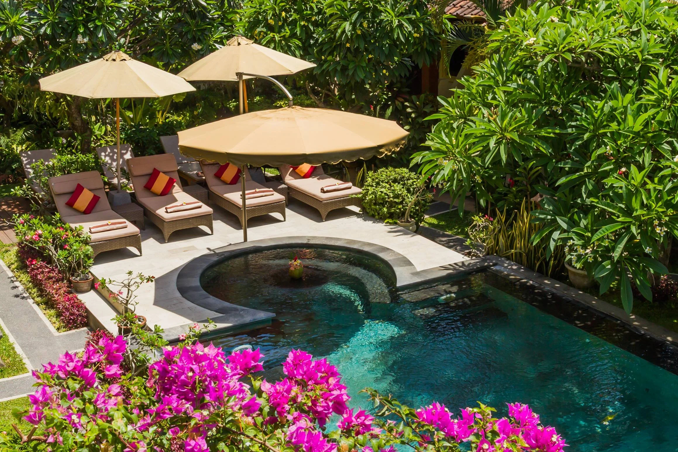 High-angle view of a secluded tropical pool surrounded by lush foliage, bright pink bougainvillea flowers, and cushioned sun loungers under shade umbrellas in Bali, photographed by Seña Studio.