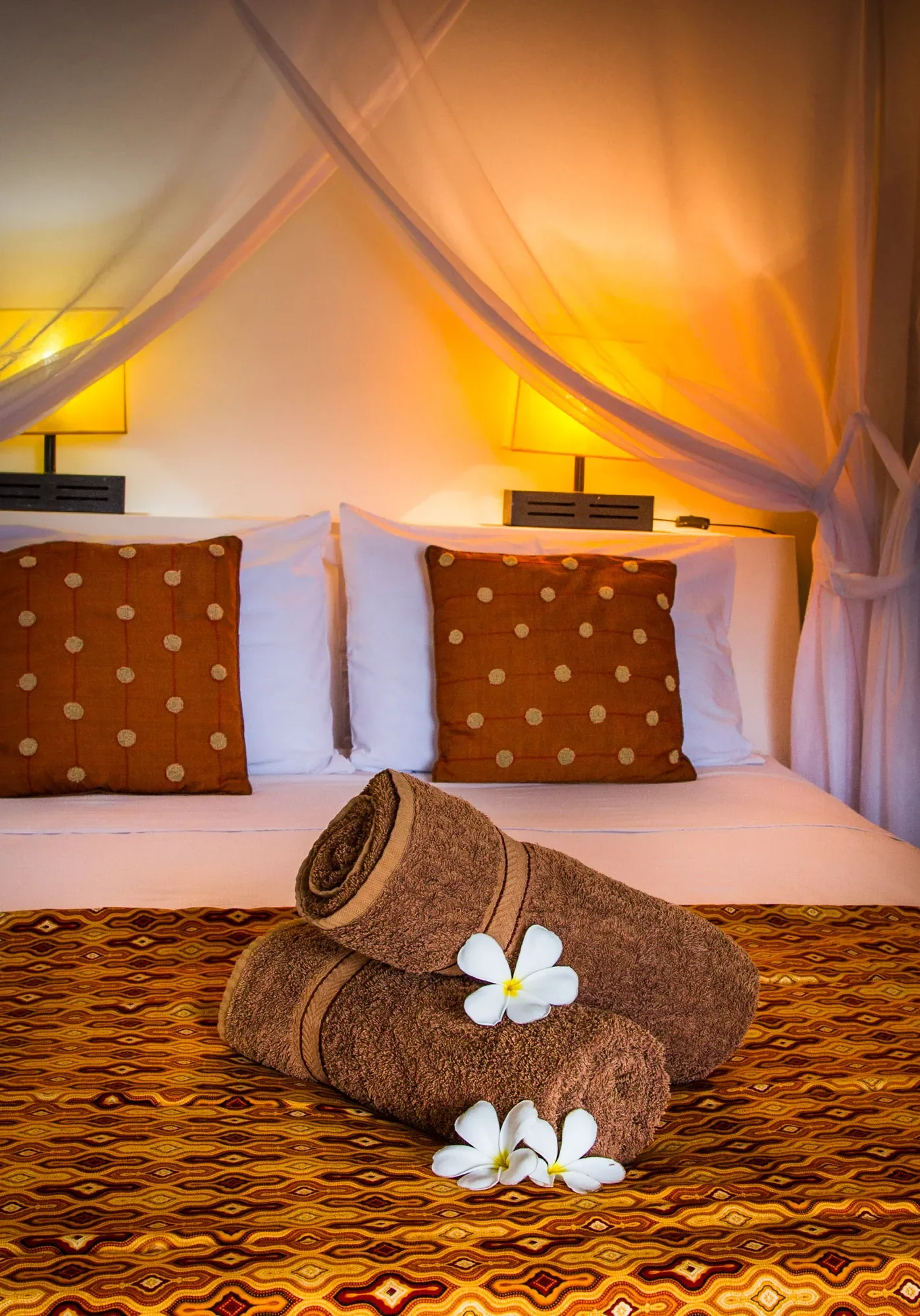 Warmly lit resort bedroom featuring a white canopy bed draped with sheer mosquito netting, accented with terracotta polka-dot throw pillows, a patterned runner, and neatly rolled brown towels topped with fresh white frangipani blossoms.