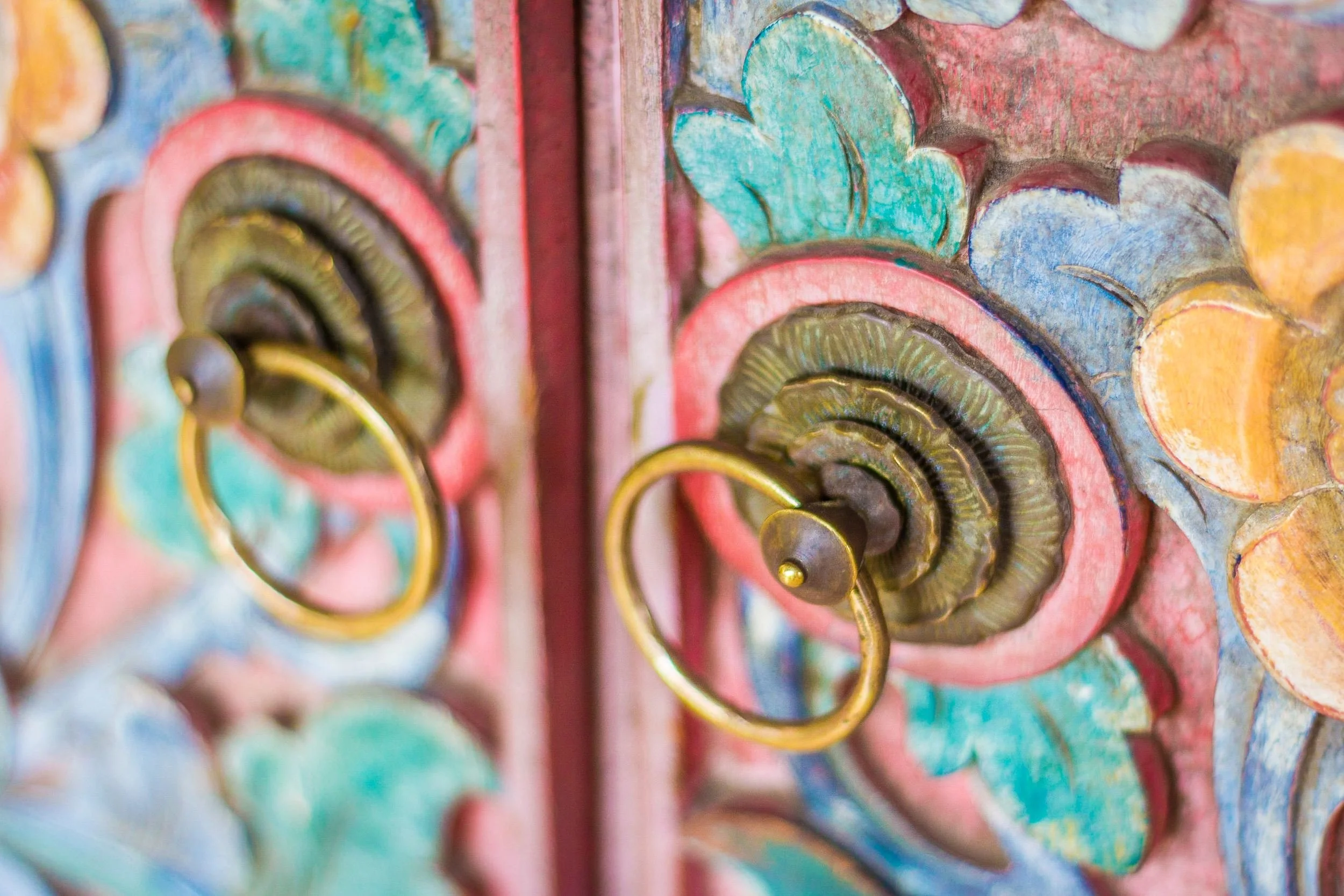 Close-up of ornate hand-carved wooden doors with brass ring handles painted in pastel turquoise and terracotta at Aquamarine Beach Villas in Amed, Bali, photographed by Seña Studio.
