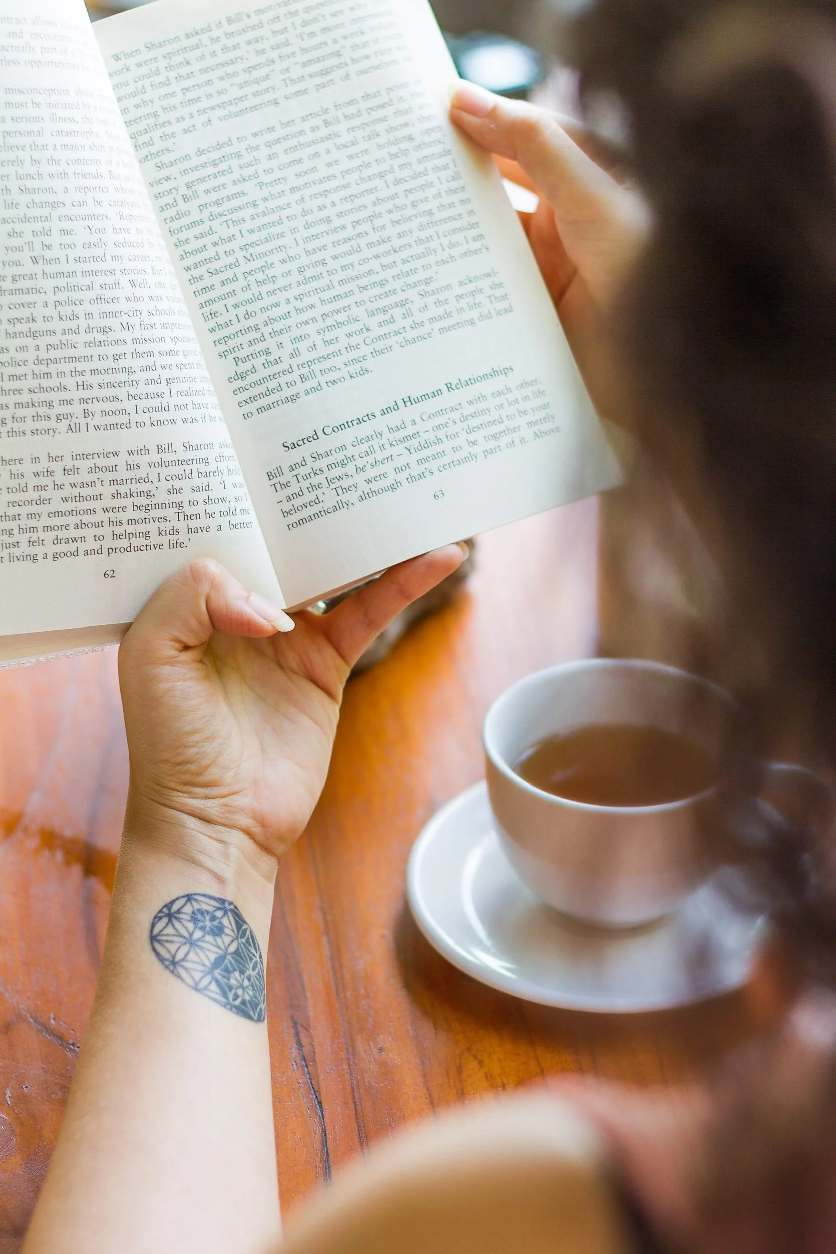 Over-the-shoulder lifestyle portrait of a person reading an open book at a warm wooden table with a steaming white cup of hot tea in the foreground, photographed by Seña Studio.