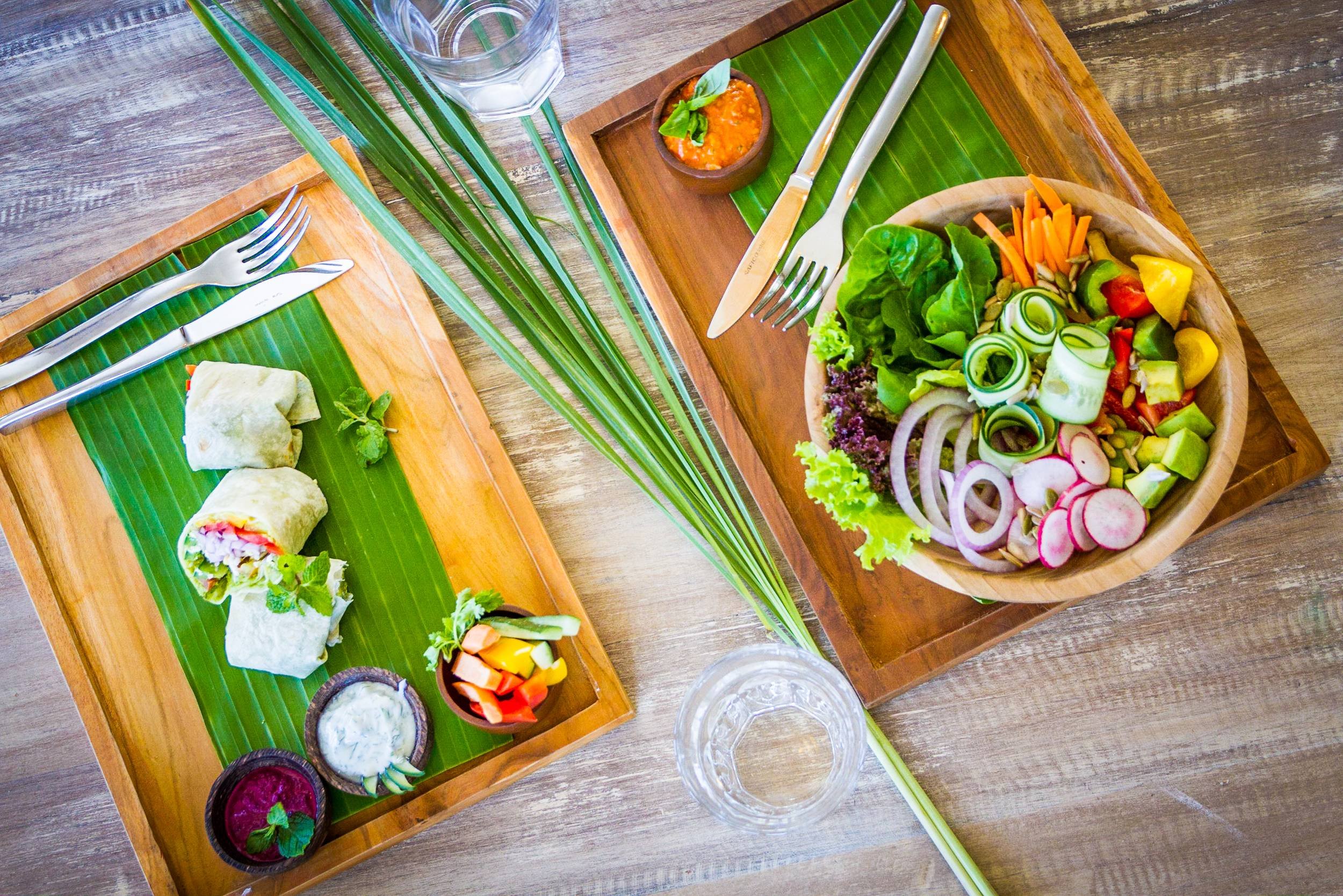 Overhead flatlay of a fresh lunch set on a wooden table with banana leaf trays, featuring cut vegetable wraps, a colorful salad bowl, dipping sauces, and glasses of water, photographed by Seña Studio.