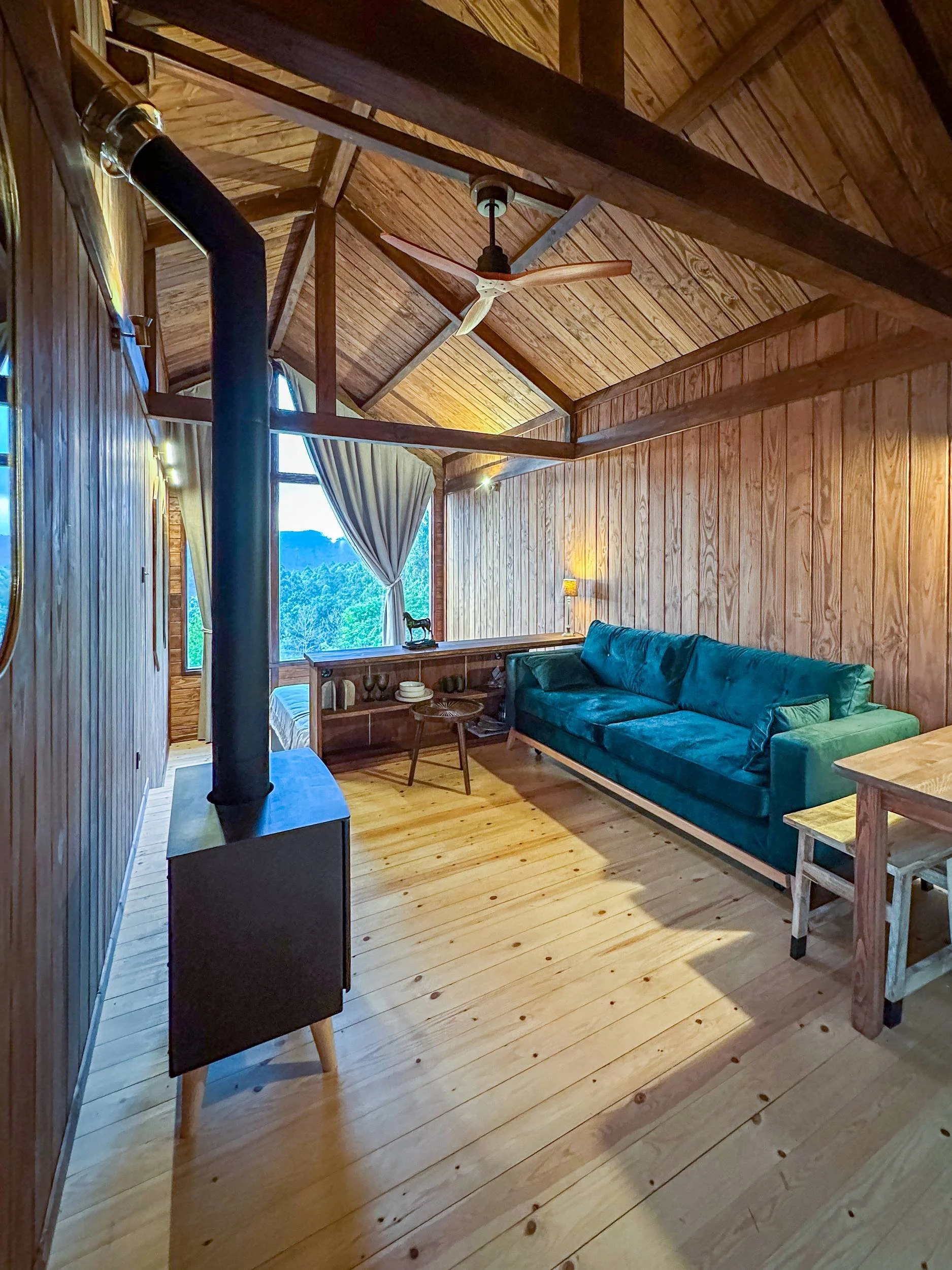 Interior view of a rustic wooden cabin living space featuring high vaulted ceilings, a black wood-burning stove, and a teal sofa at Haras de Bordo, photographed by Seña Studio.