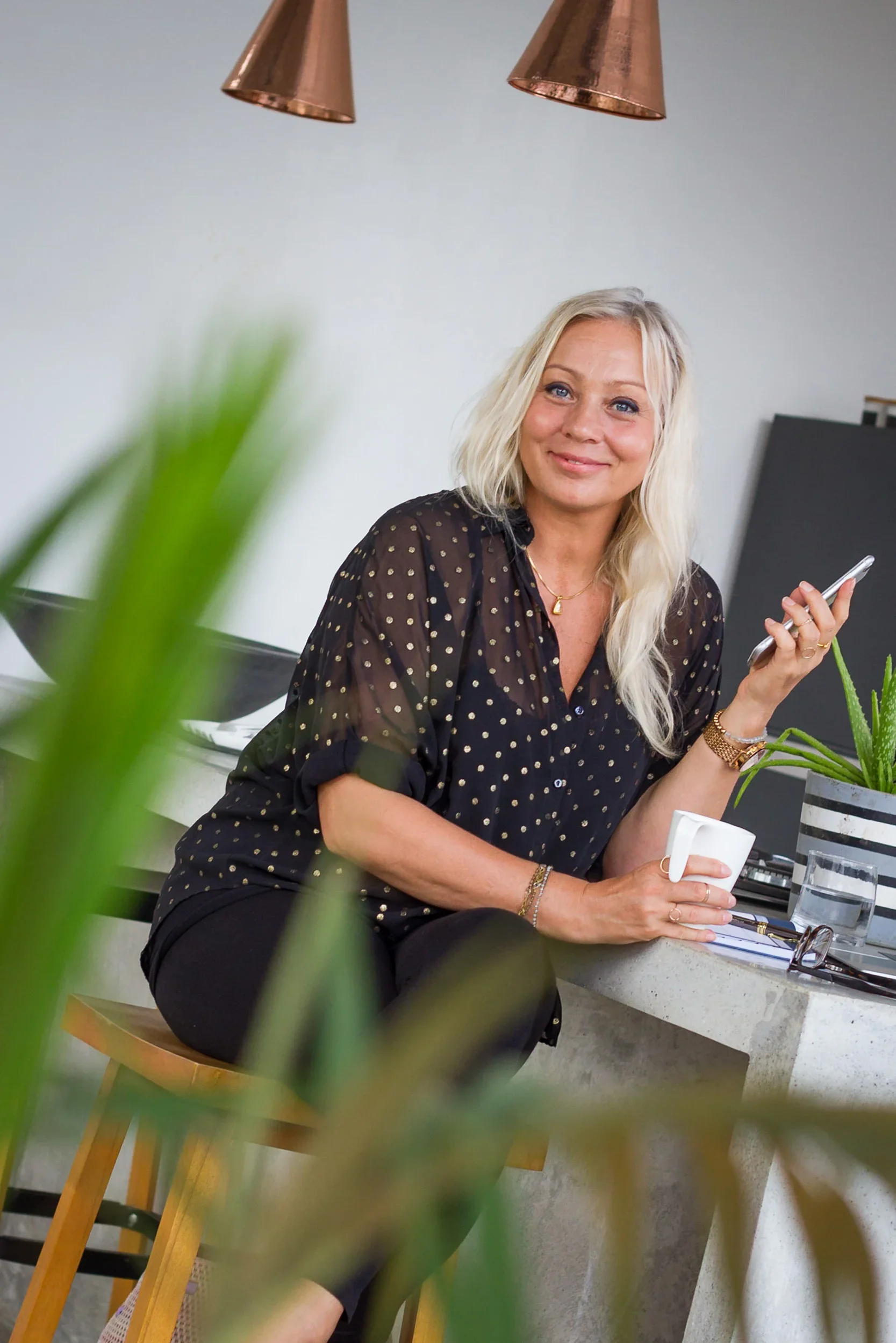 Female entrepreneur in a black-and-gold polka-dot blouse resting at a counter with a coffee mug and a smartphone.