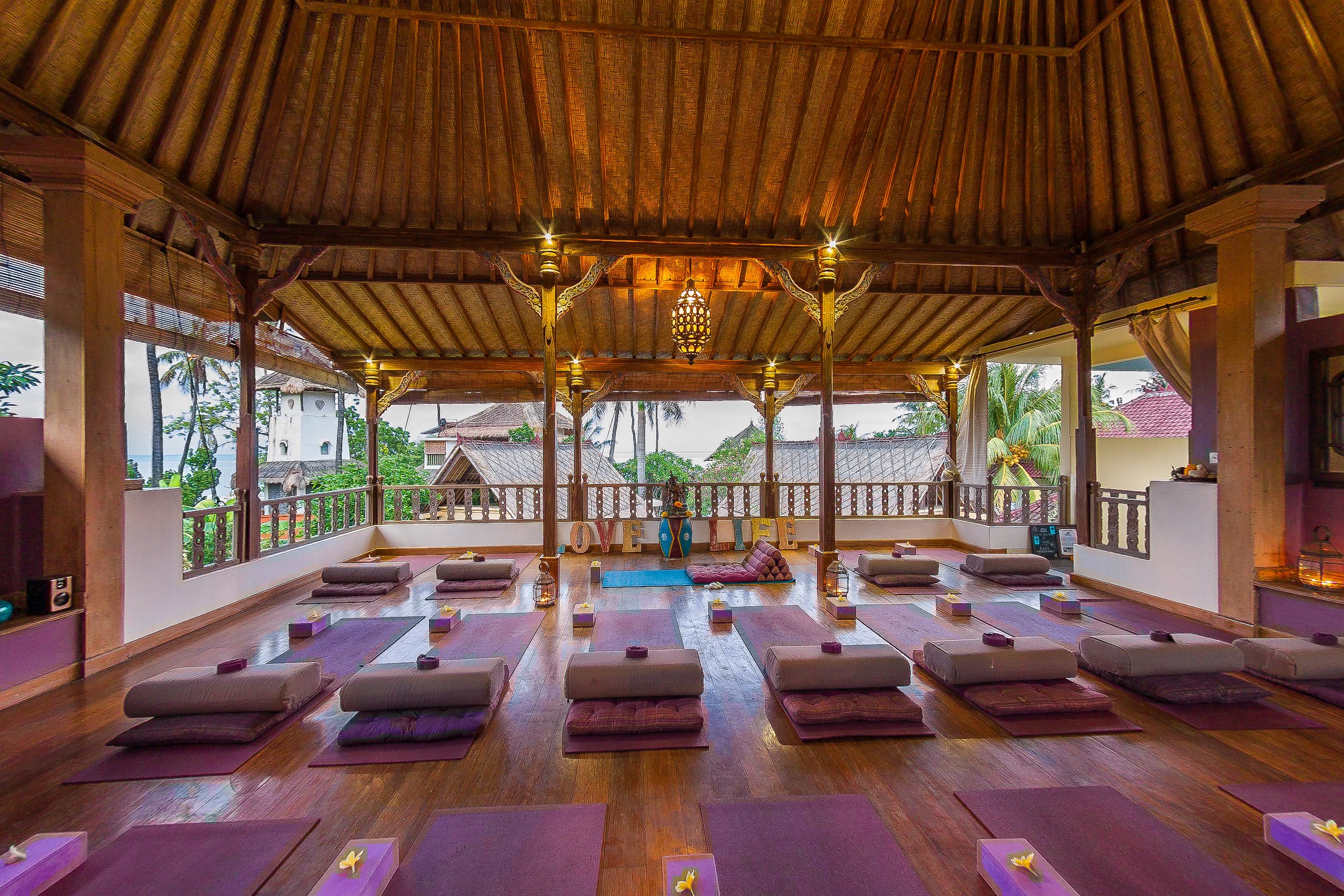 Wide-angle shot of a spacious open-air wooden yoga shala with intricate carved pillars, thatched roof, and neatly arranged purple yoga mats with bolsters, photographed by Seña Studio.