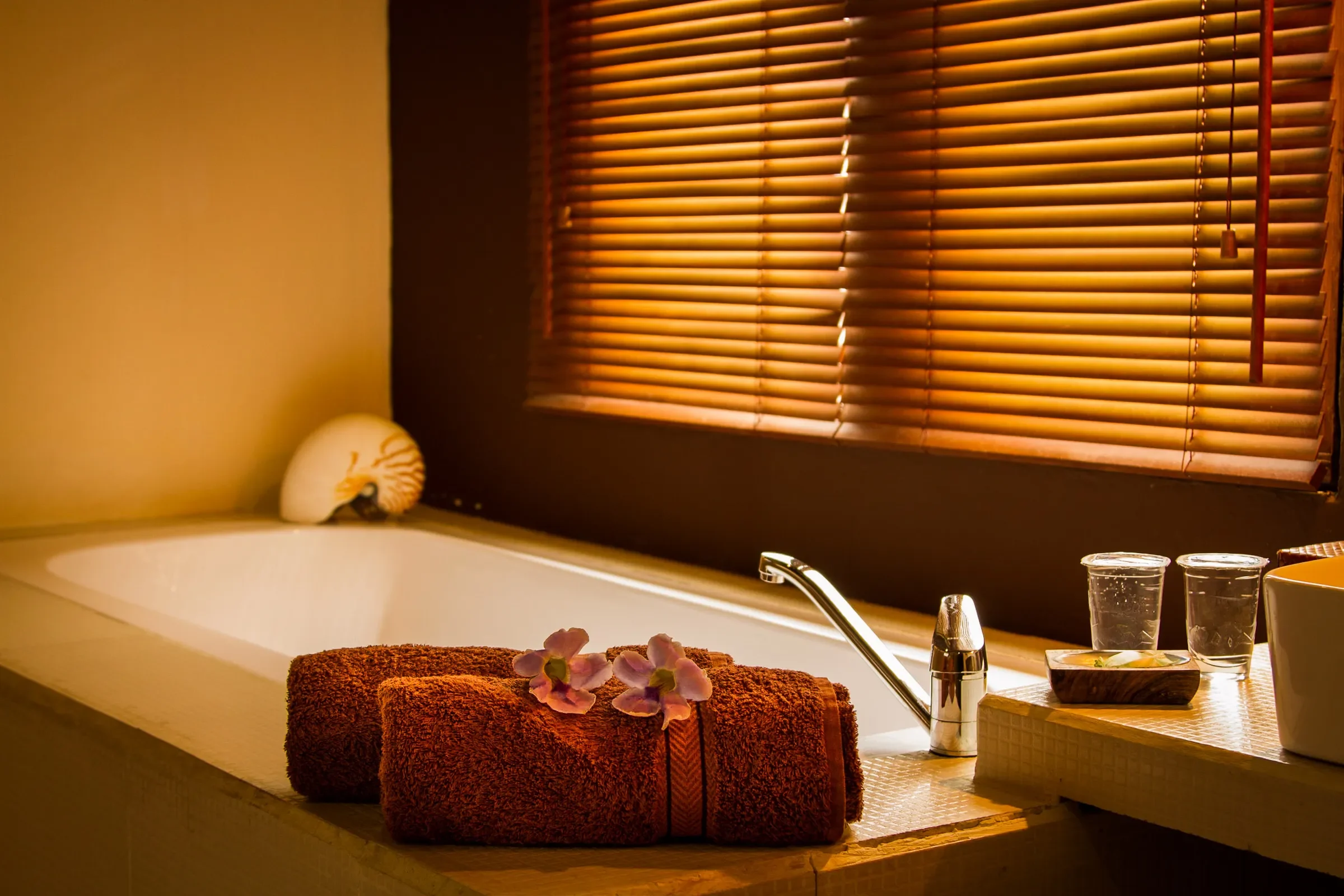 An intimate and warm spa-style bathroom interior view, showing a corner of a white built-in bathtub. In the foreground, two neatly rolled terracotta-orange towels are adorned with delicate purple flowers. A Nautilus shell sits on the ledge, and golde