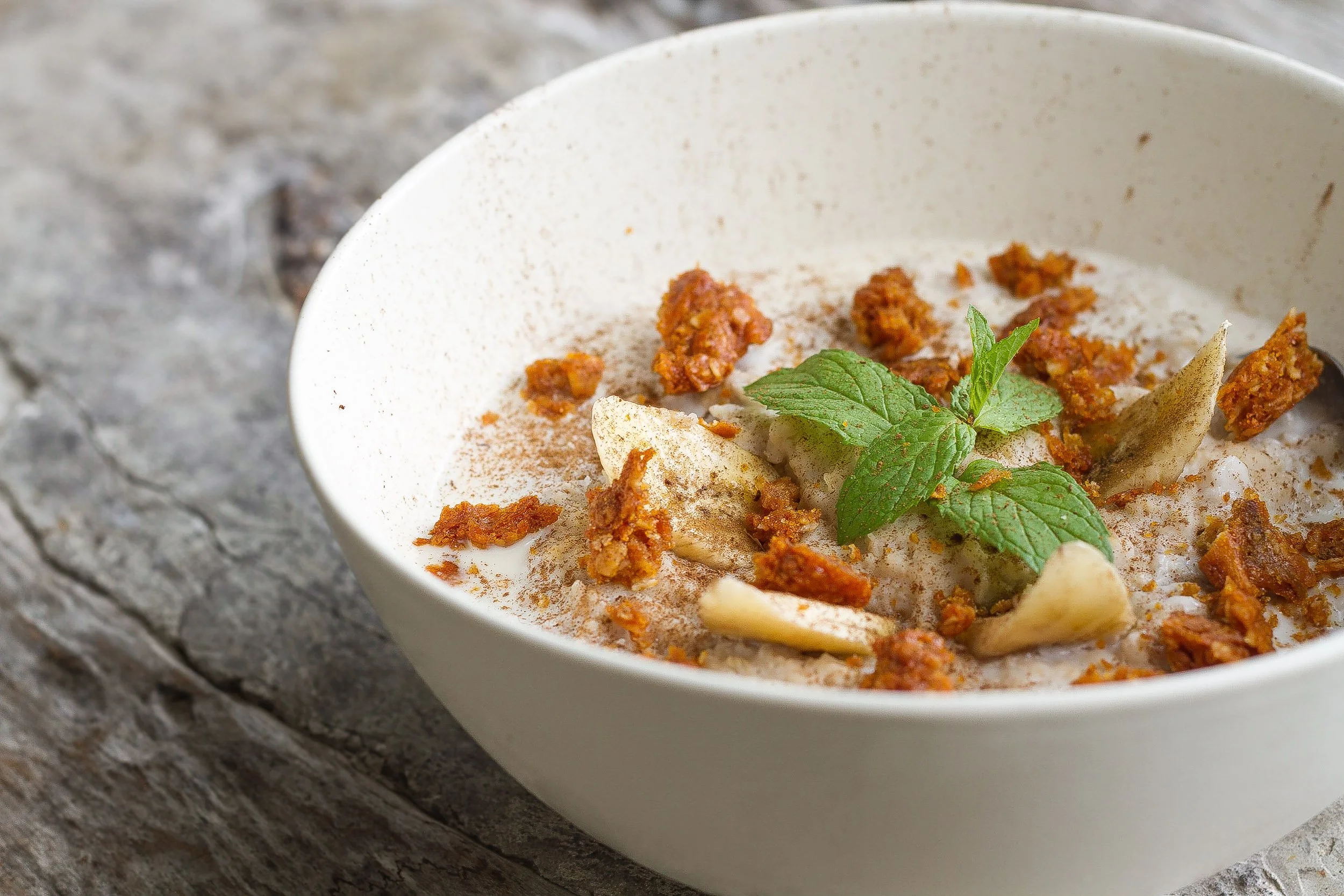 A close-up shot of a white bowl featuring oatmeal topped with banana slices, crunchy granola clusters, dusted cinnamon, and a sprig of fresh mint set on a rustic wooden table.