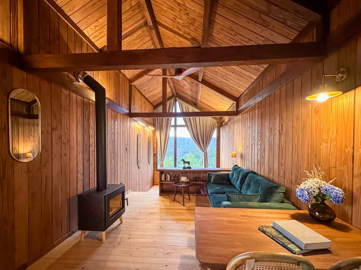 Warm wooden cabin interior with vaulted ceilings, a wood-burning stove, green velvet sofa, and large picture window at Haras de Bordo, photographed by Seña Studio.