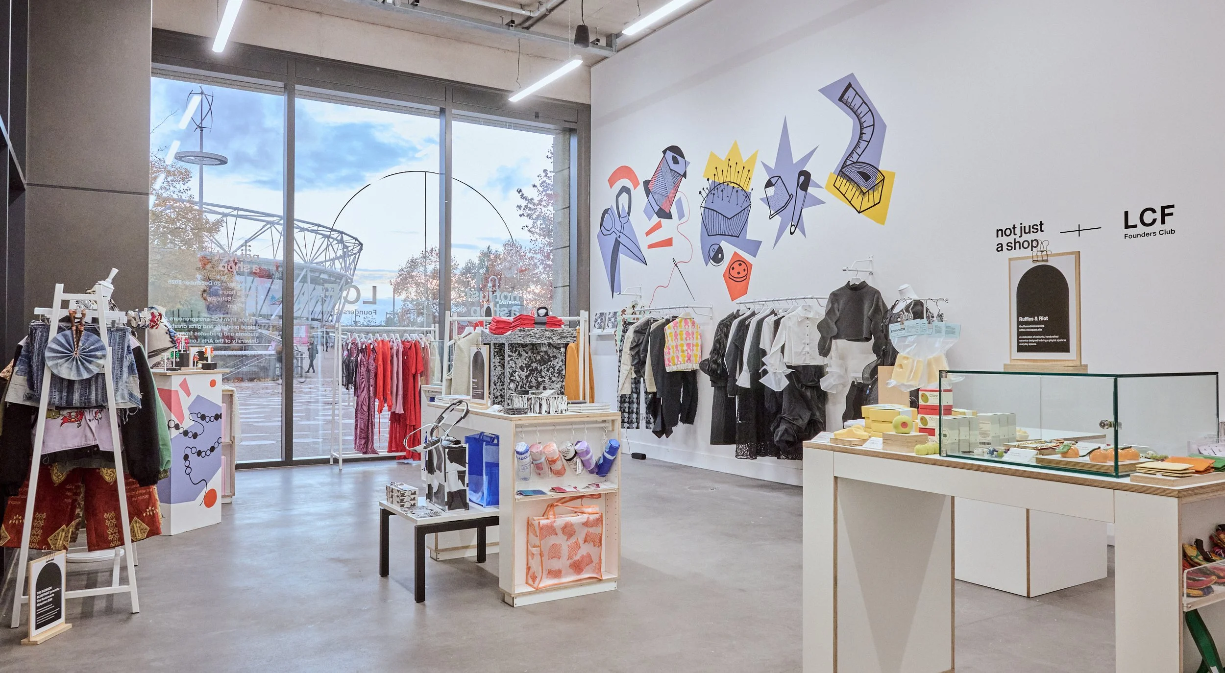 Interior of a modern store with colorful wall art, clothing racks, display tables, and a large glass window showing an outdoor stadium structure.