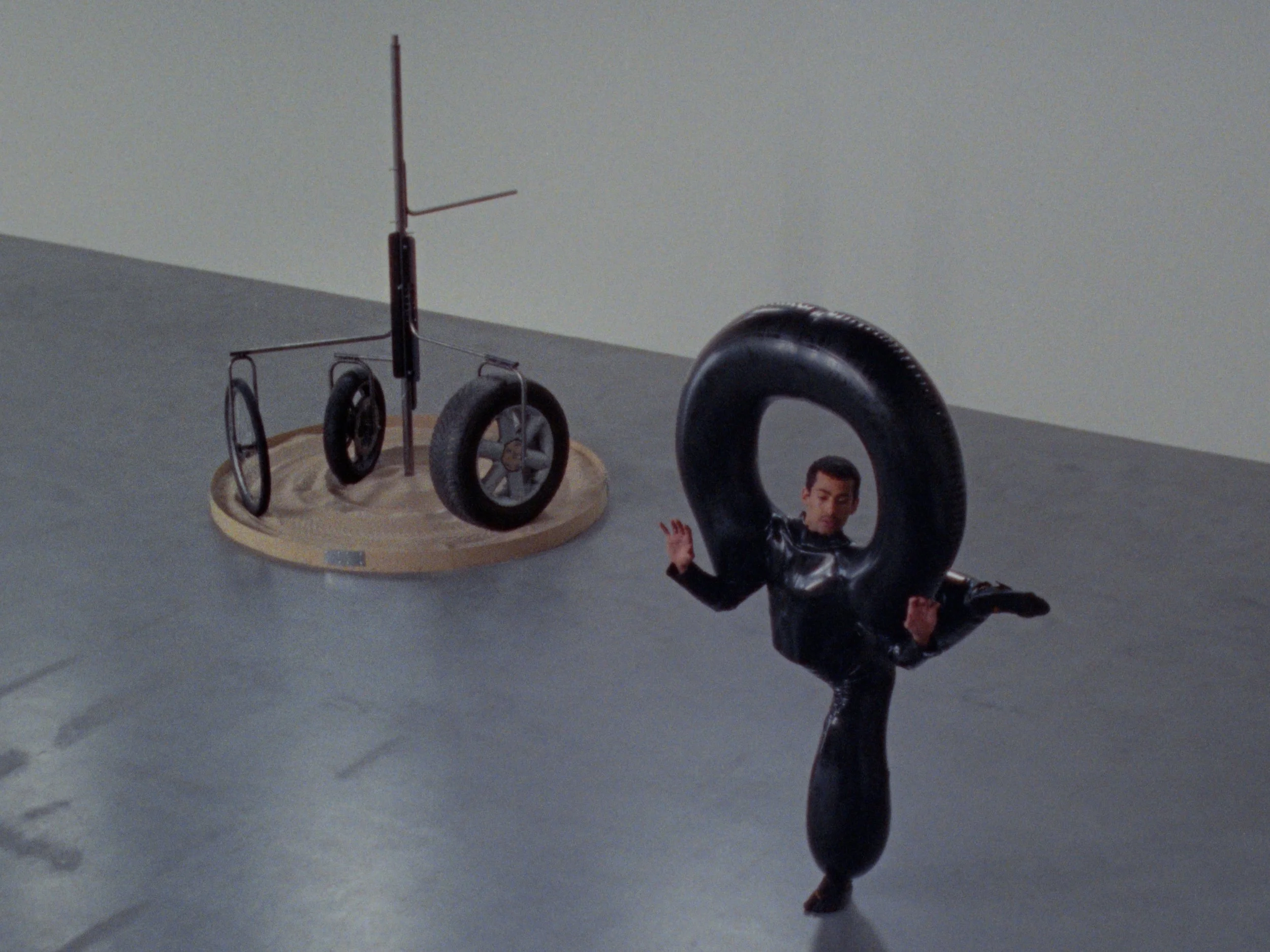 A man in a black, inflatable avant-garde outfit balances on one leg whilst standing next to a sculpture made of rubber tires and wood.