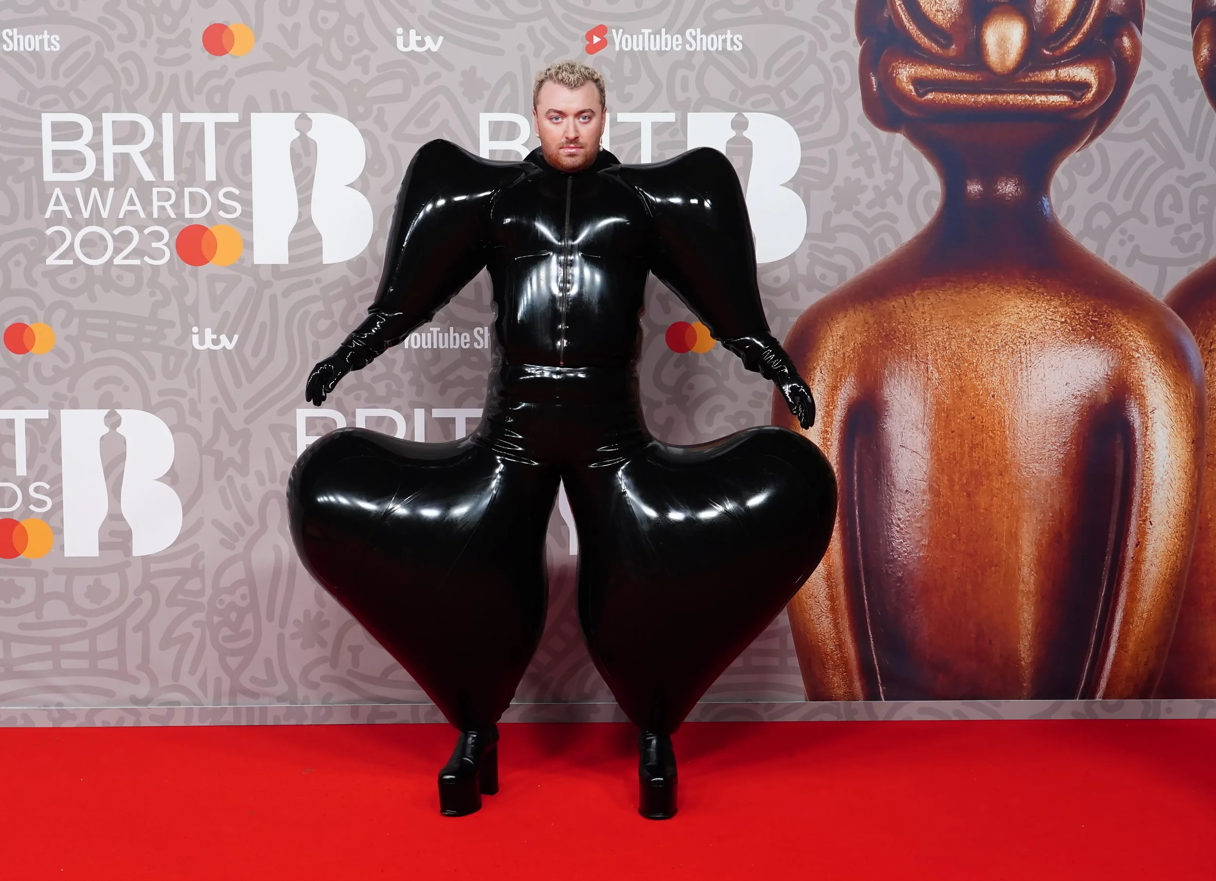 A bold person wears a shiny black avant-garde outfit with exaggerated shoulders and sculpture-like pants, posing on a red carpet at the 2023 BRIT Awards event.