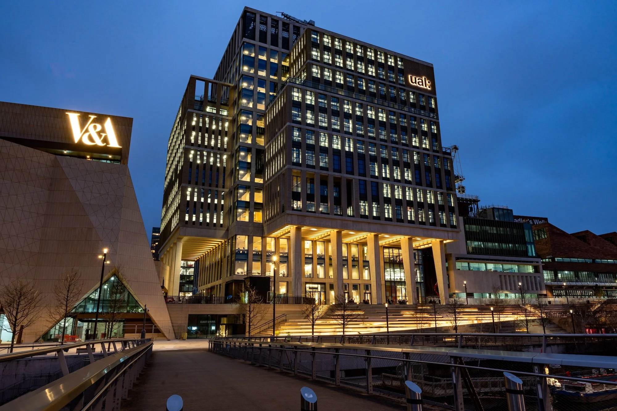 Night view of modern city buildings with illuminated windows, including a high-rise with signage 'V&A' and 'UAL', and an architectural structure with geometric design, along a waterfront walkway with leafless trees and street lamps.