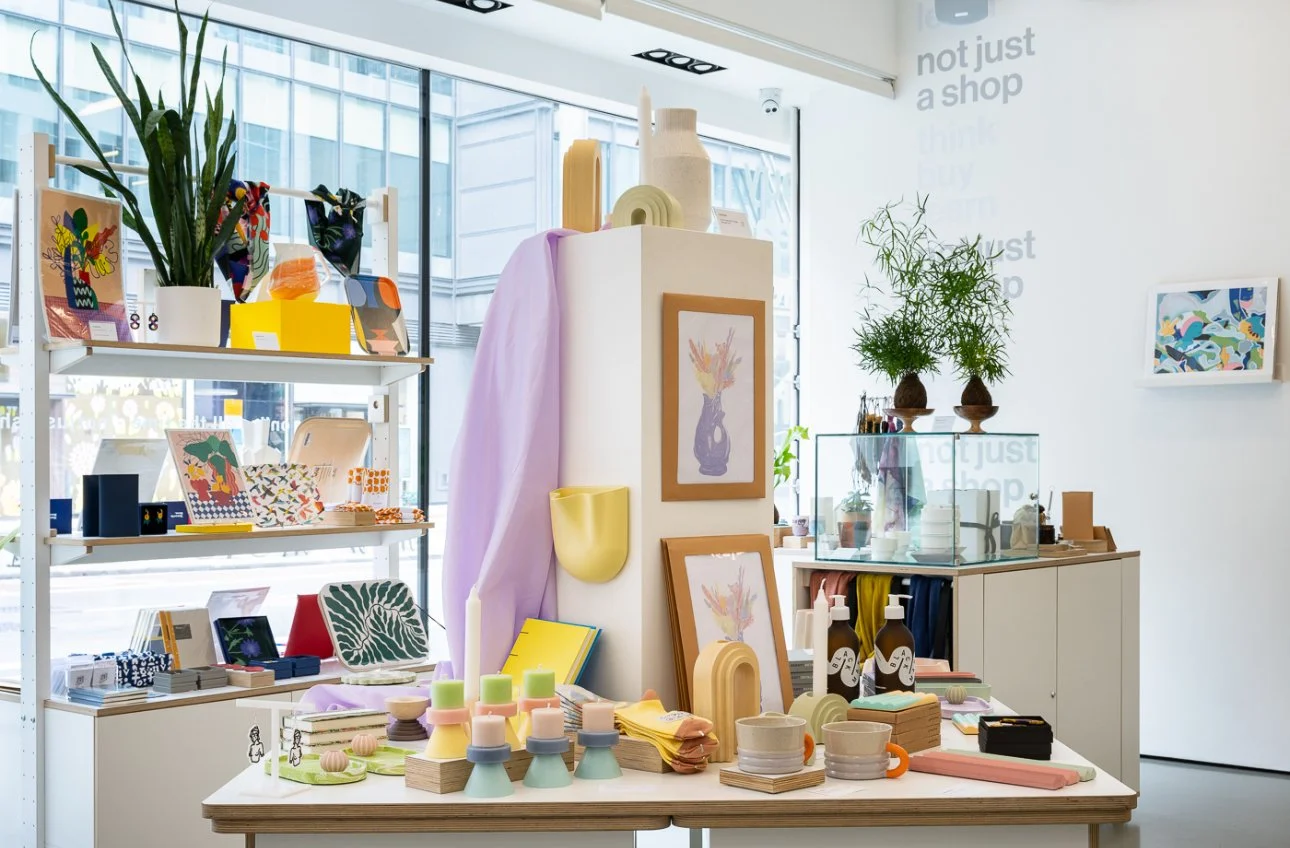 Interior of a boutique shop with shelves of colorful home decor items, framed artwork, potted plants, and pastel-colored accessories displayed on a table and shelves.
