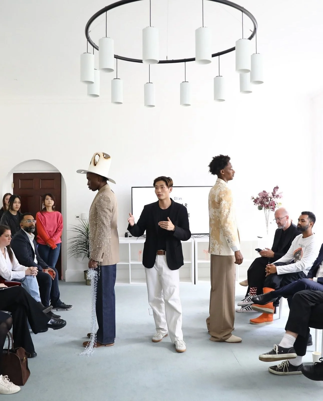 A presentation is taking place in a bright, white-walled room with a chandelier and a large monitor. There are multiple people seated and standing, some dressed fashionably, with three models showcasing clothing in the center.