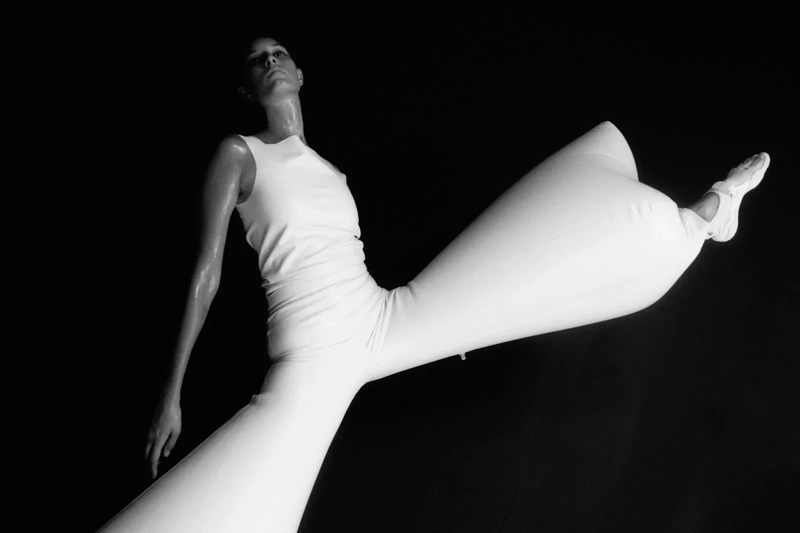 A female dancer elongates her leg into the air whilst wearing a white sculpture against a black background, backstage during London Fashion Week.