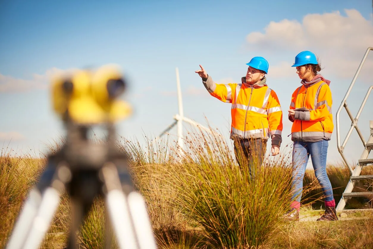 Two construction workers in orange safety jackets and blue helmets working outdoors near wind turbines, with surveying equipment in the foreground.