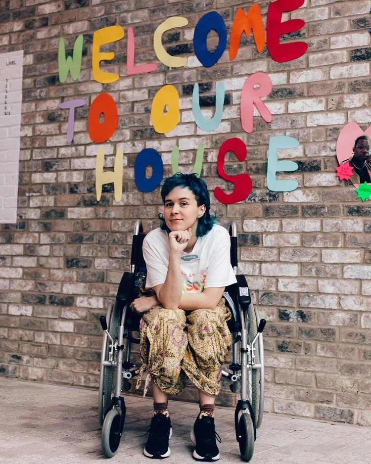 A disabled non-binary dancer sits in a wheelchair in front of a brick wall with colourful paper letters that spell 'Welcome to Our House'. 