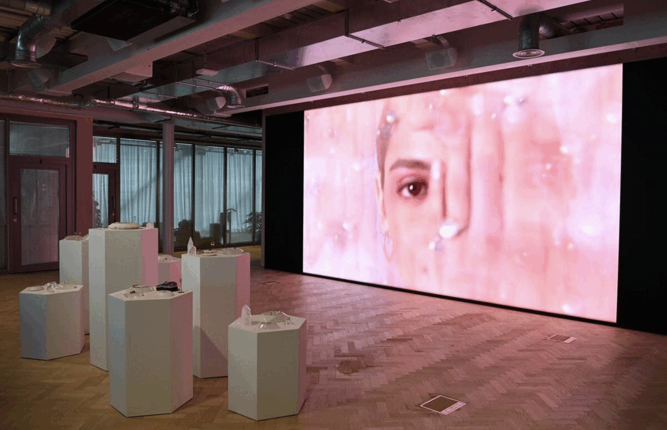 A contemporary wellness space with a large digital screen displaying a close-up of a person's face with a pink background. There are six white pedestal tables arranged in front of the screen, each with delicate fine jewellery crystals.