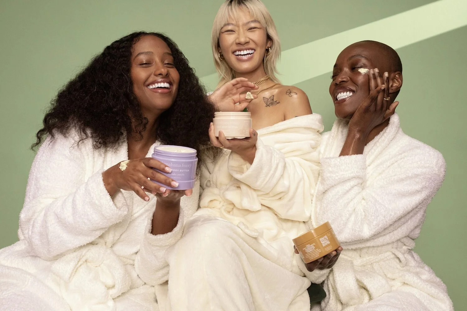 Three women in white robes smiling and laughing together, holding containers of body butter, against a light green background.