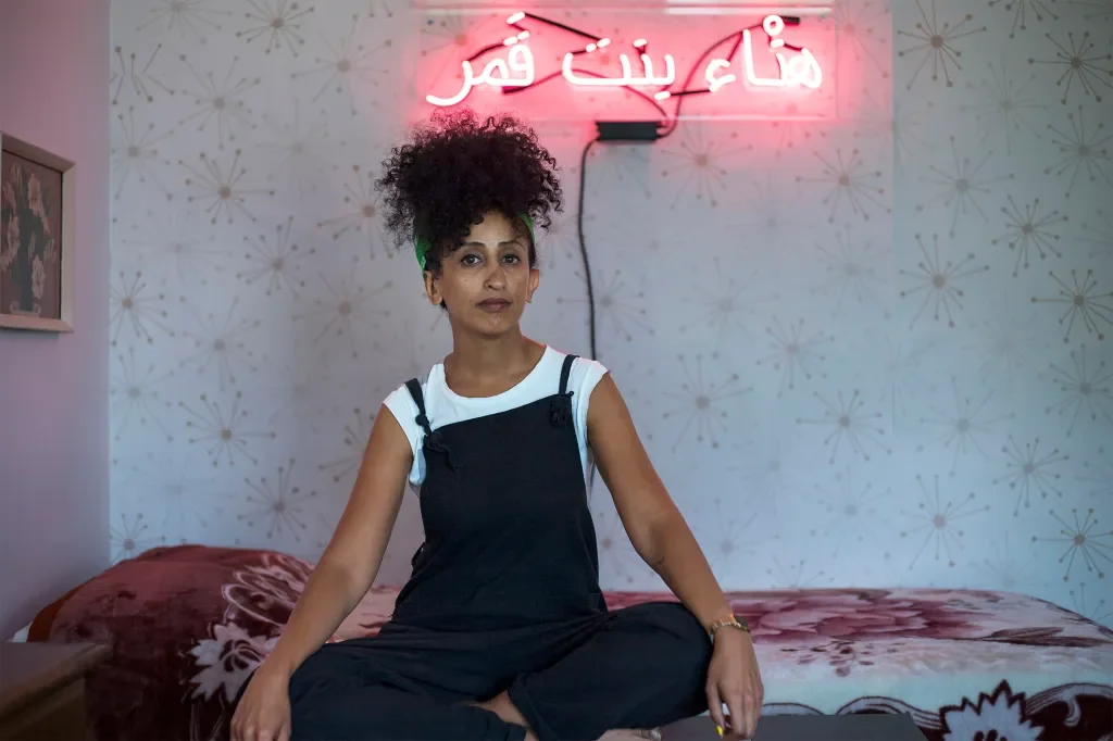 A woman with curly hair tied up with a green headband, sitting cross-legged on a bed with floral bedding in a room with a pink neon sign in Arabic and patterned wallpaper.