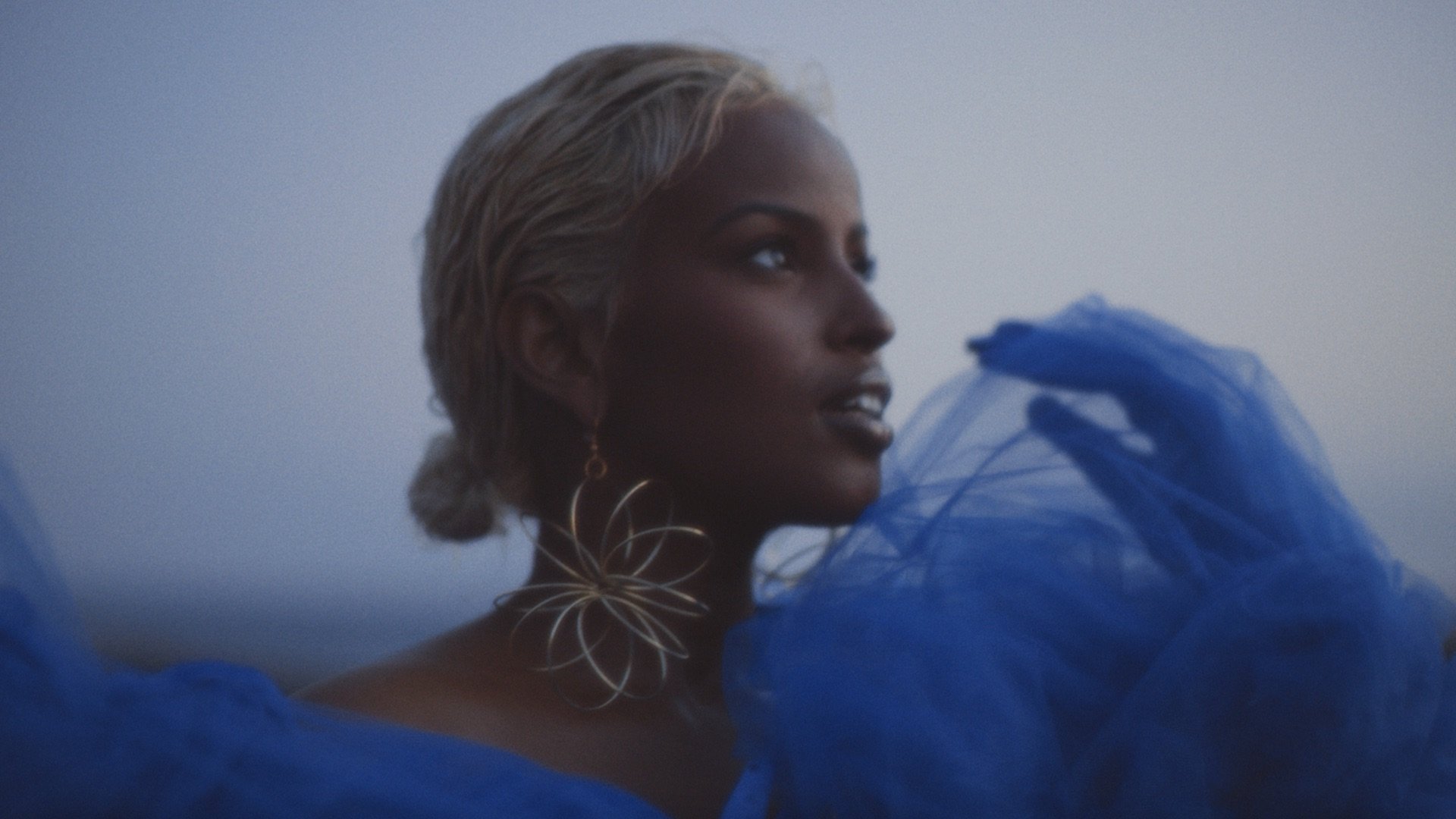 A woman with blonde hair styled in an updo, wearing large floral-shaped earrings and a matching floral choker necklace, draped in blue fabric.