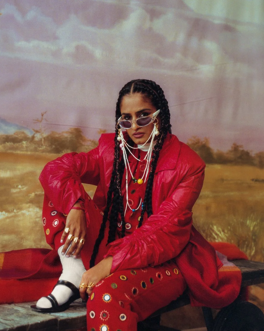 A woman with long braided hair wearing sunglasses, multiple necklaces, gold rings, a red jacket, and patterned pants, sitting outdoors against a nature backdrop.