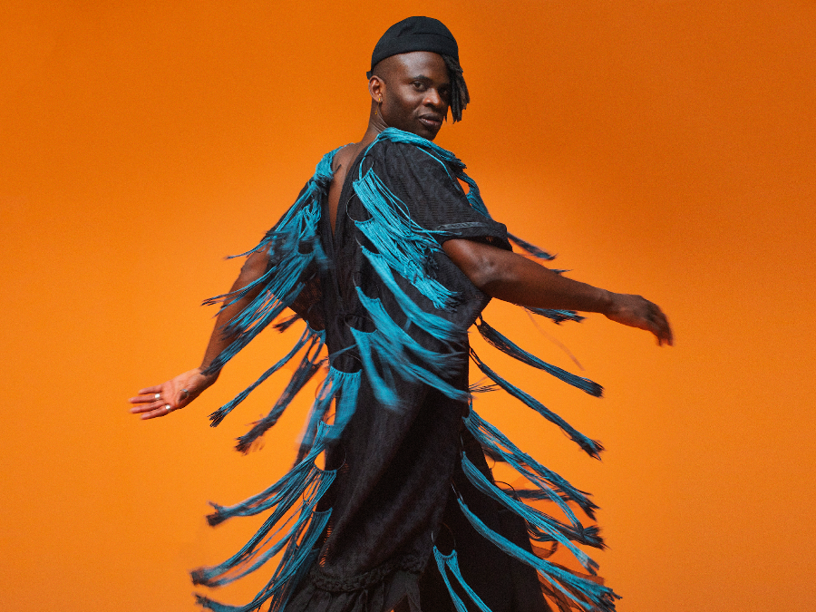 A male musicians wears a black outfit with blue fringes, posing against an orange background.