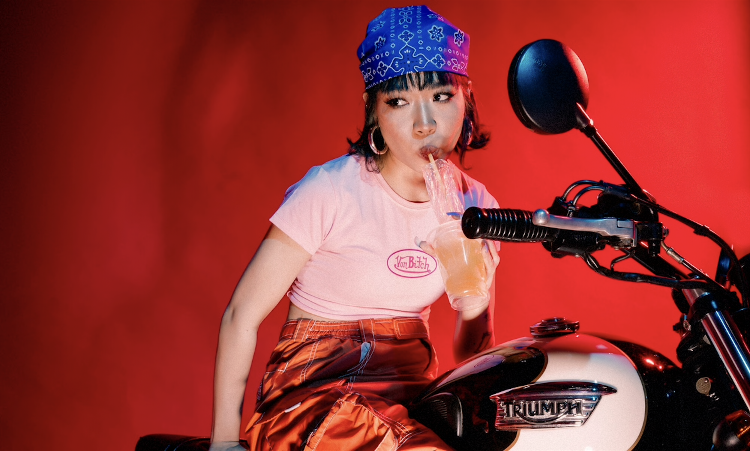 A woman with a blue bandana drinking a beverage with a straw from a clear plastic cup while sitting on a Triumph motorcycle with a red background.