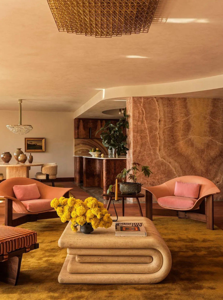 Layered living room with coral lounge chairs, a sculptural stone coffee table, dramatic rose-colored stone walls, warm wood, and collected decorative pieces.