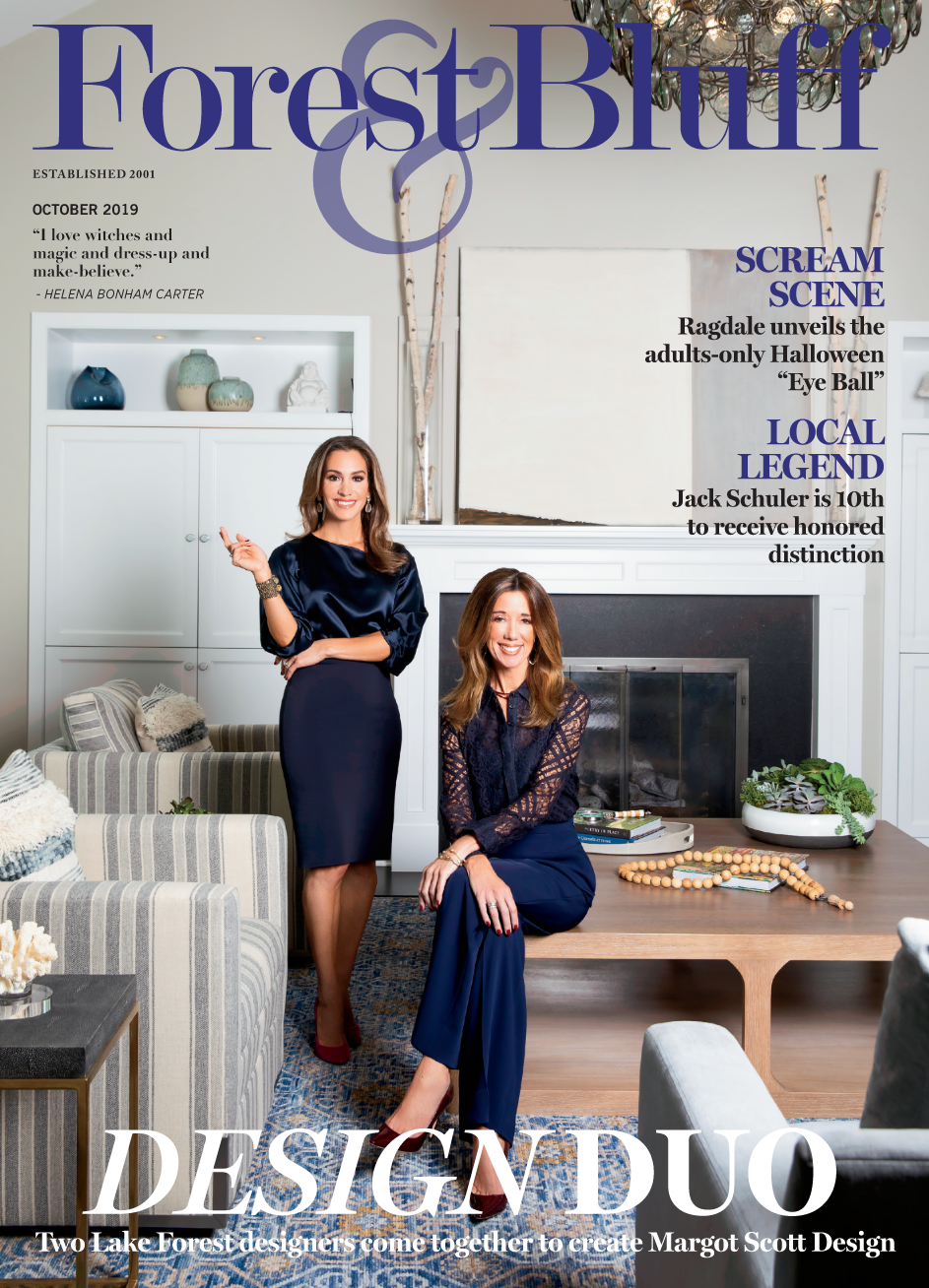 Cover of Forest & Bluff magazine featuring two women in a living room, one standing and one sitting on a table, with a fireplace and decorative items in the background at home.