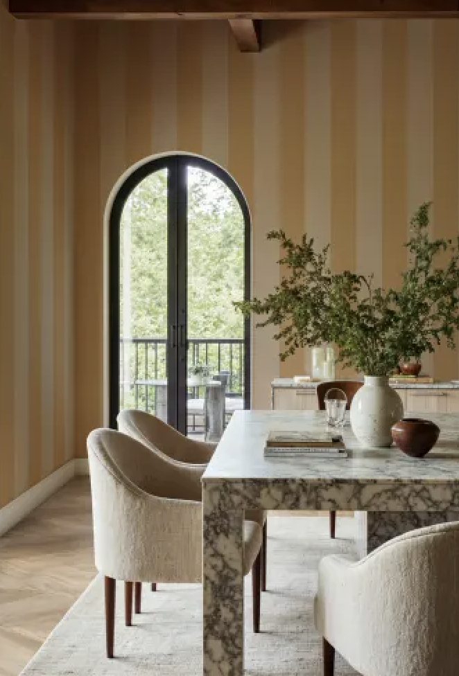 A dining room with marble stone table, yellow ochre striped wallpaper. white bucket dining chairs, and greenery in a vase with a scenic view.
