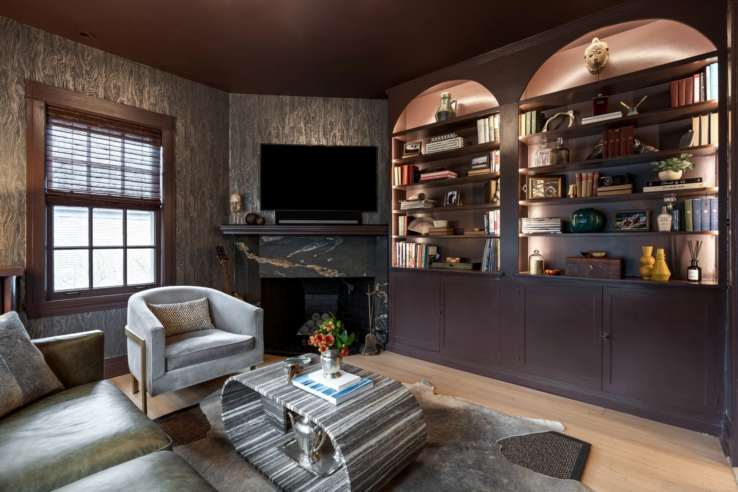Moody office lounge space with custom bookshelves builtins, metallic woodlike wallpaper, layered cowhide rug, unique stone coffee table with orange flowers, books and fireplace.