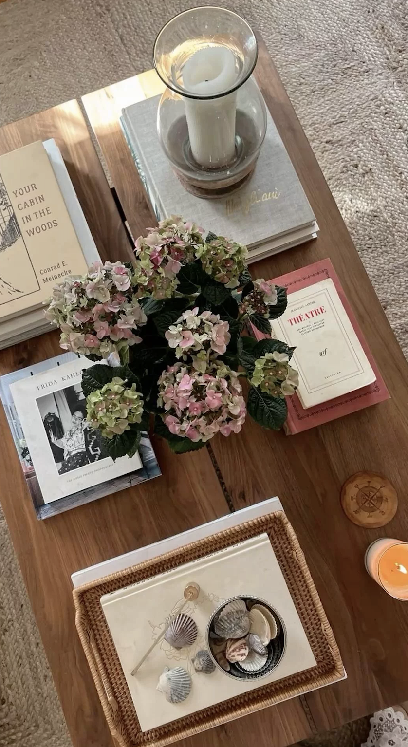 Wood coffee table styled with hydrangeas, books, seashells, woven accessories, and softly glowing candles.