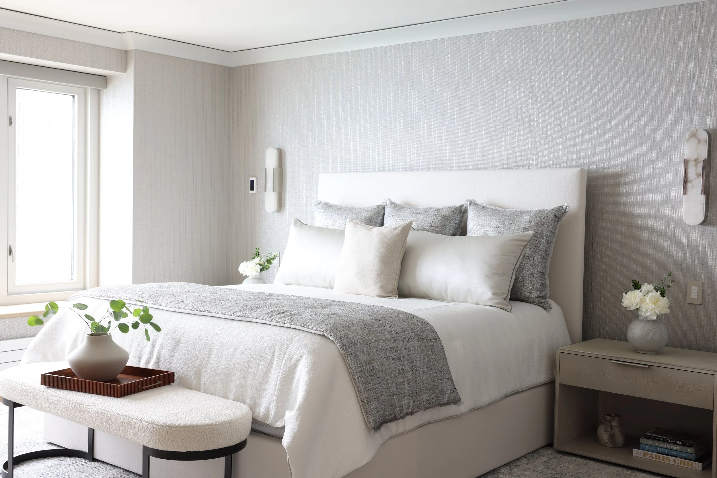 A modern bedroom with white walls, a large bed with white and gray pillows, a white headboard, a bedside table with a vase of white flowers, and a window with white blinds.