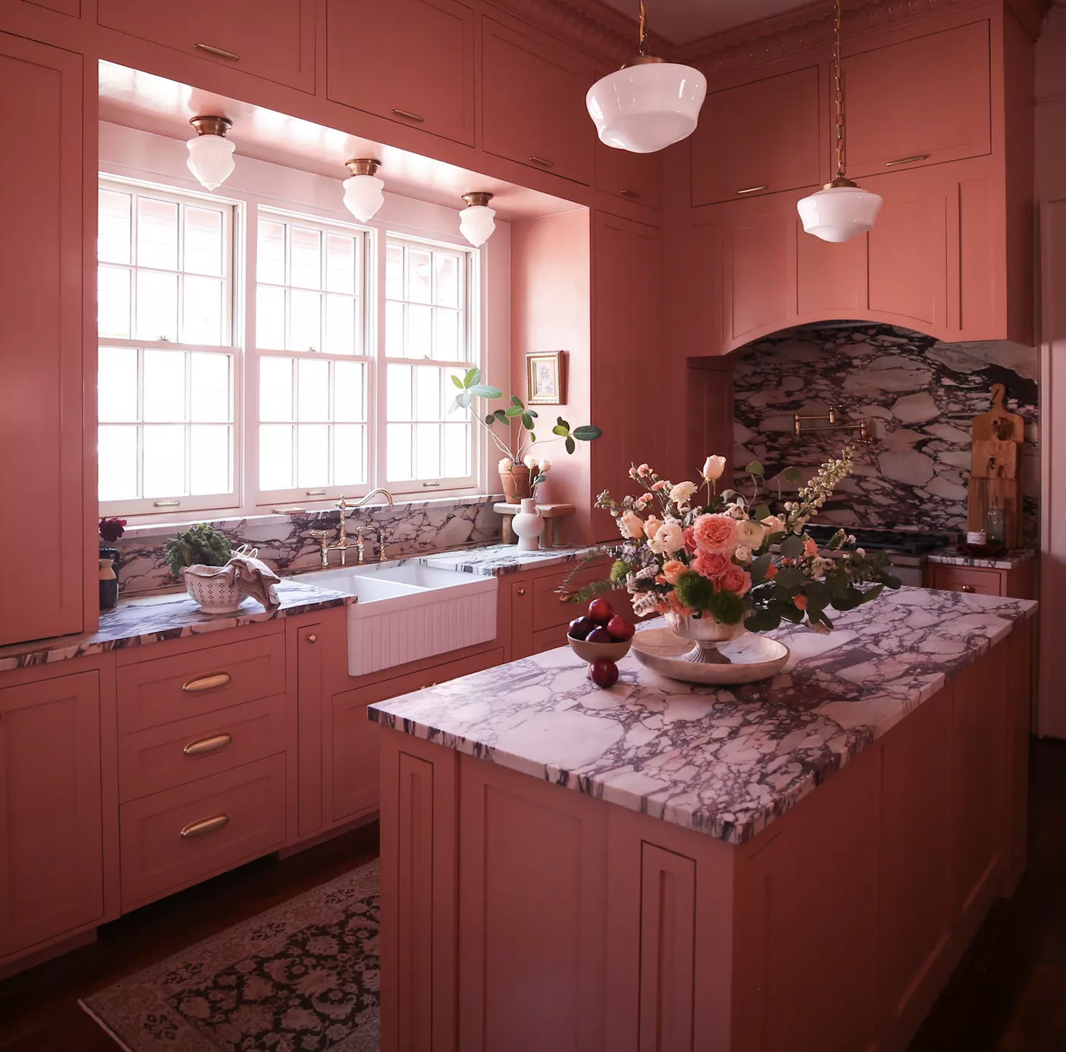 Dramatic color drenched pink kitchen with custom paneled cabinetry, brass hardware, vintage-inspired lighting, a farmhouse sink, and richly veined marble countertops and backsplash.
