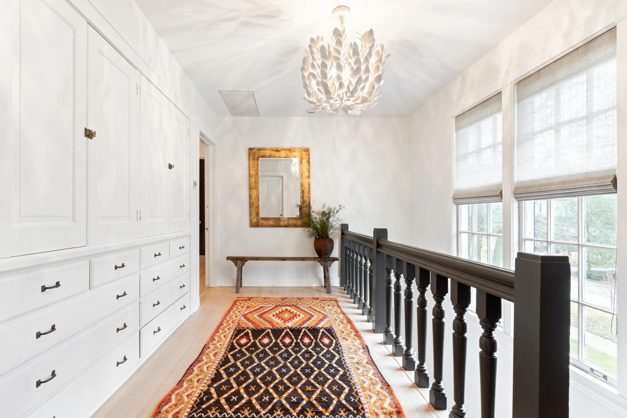 Interior view of a bright upstairs landing with white walls, a decorative black railing, large windows with blinds, a gold framed mirror, a wooden bench, a potted plant, a colorful ethnic patterned rug, and a coconut shell chandelier.