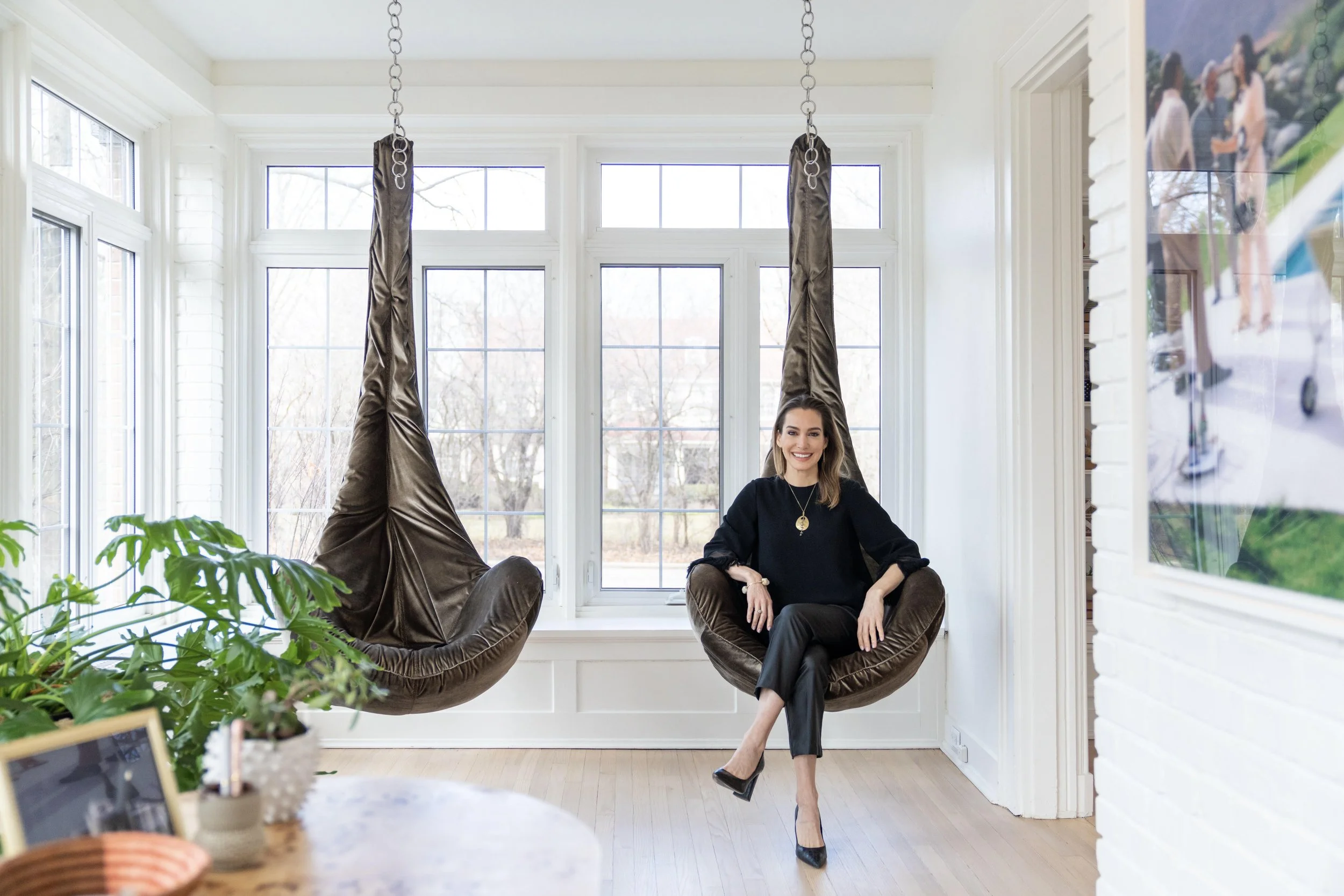 Woman sitting in a brown hanging chair in a bright room with large windows, smiling, with greenery and a blurred photo of friends in the background.