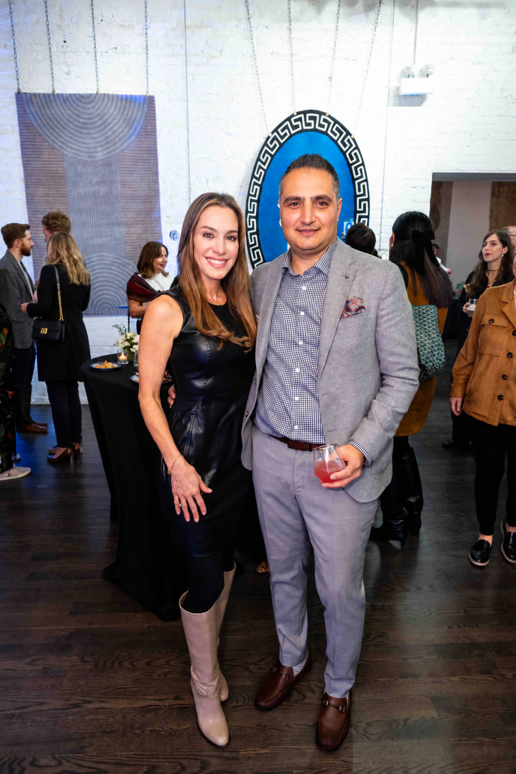 A woman in a black leather dress and beige knee-high boots standing next to a man in a light gray suit and checkered shirt, holding a glass of drink at an indoor social event.