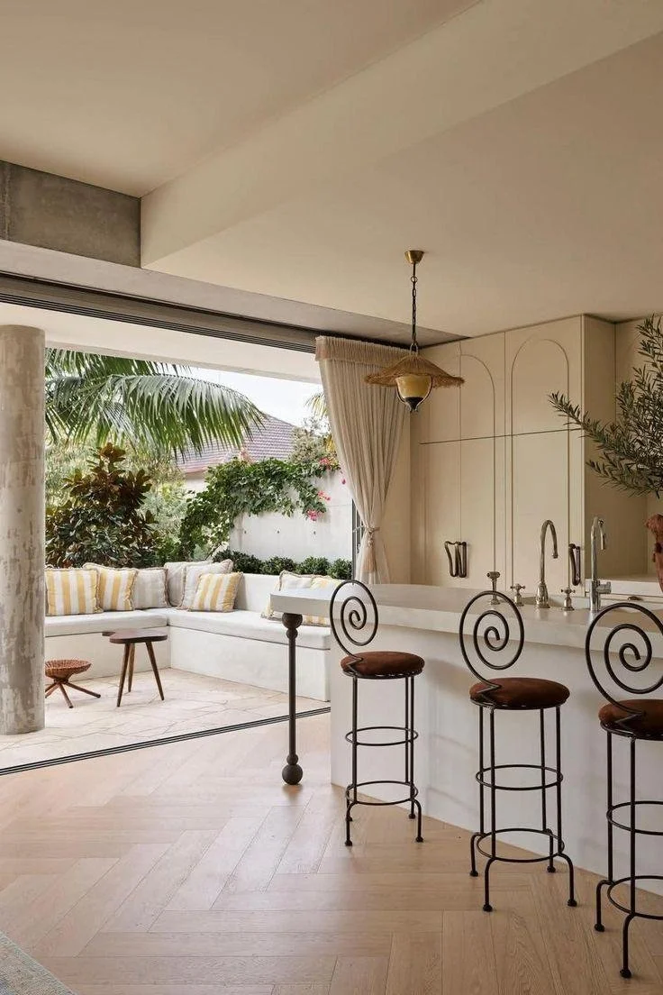Open-air kitchen with sculptural barstools, natural wood flooring, tropical greenery, and a relaxed outdoor seating area.