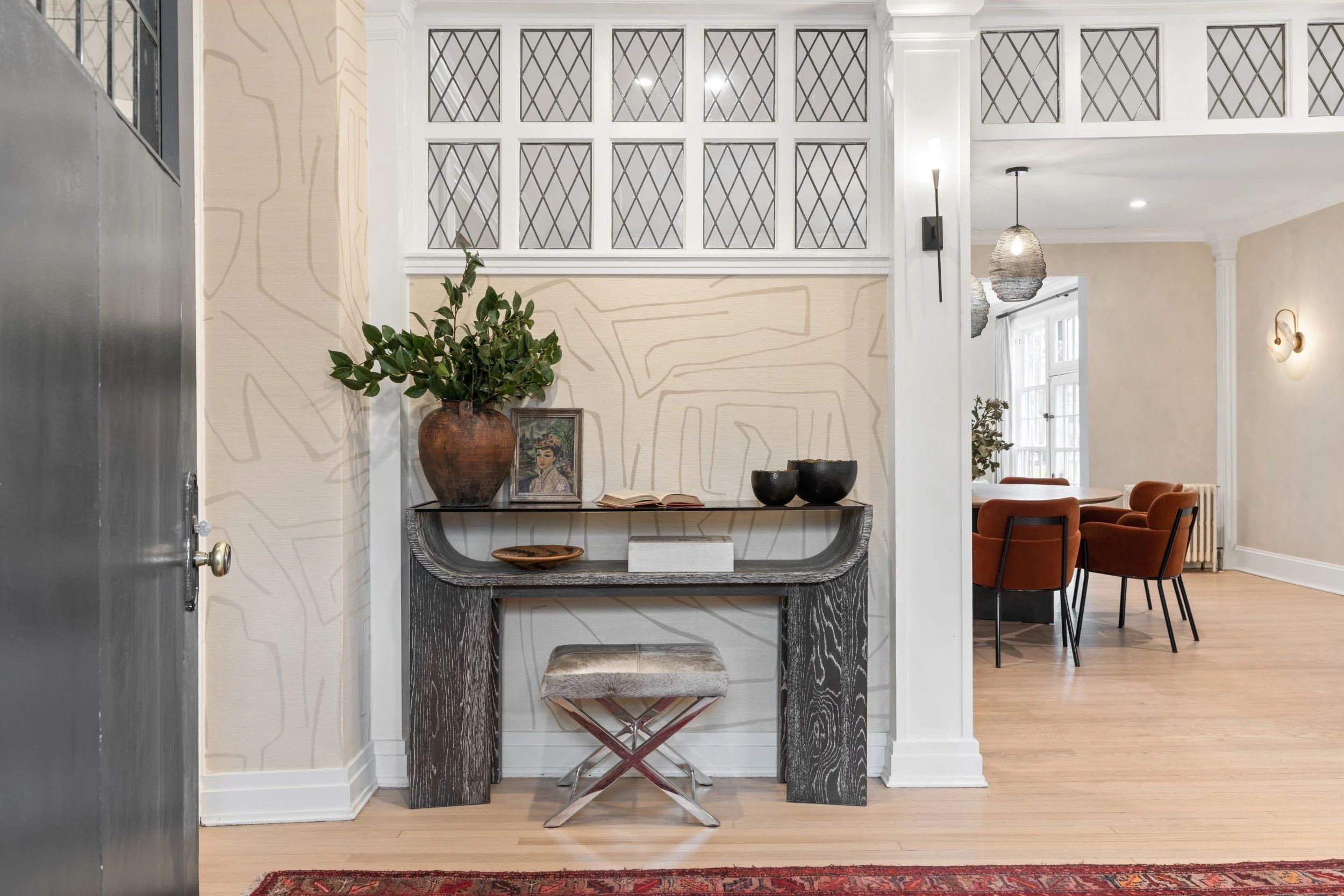 Interior view of a entryway with a console table, bold wallpaper, green potted plant, framed artwork, and decorative bowls. The hallway opens into a dining area with orange rust chairs and large windows.
